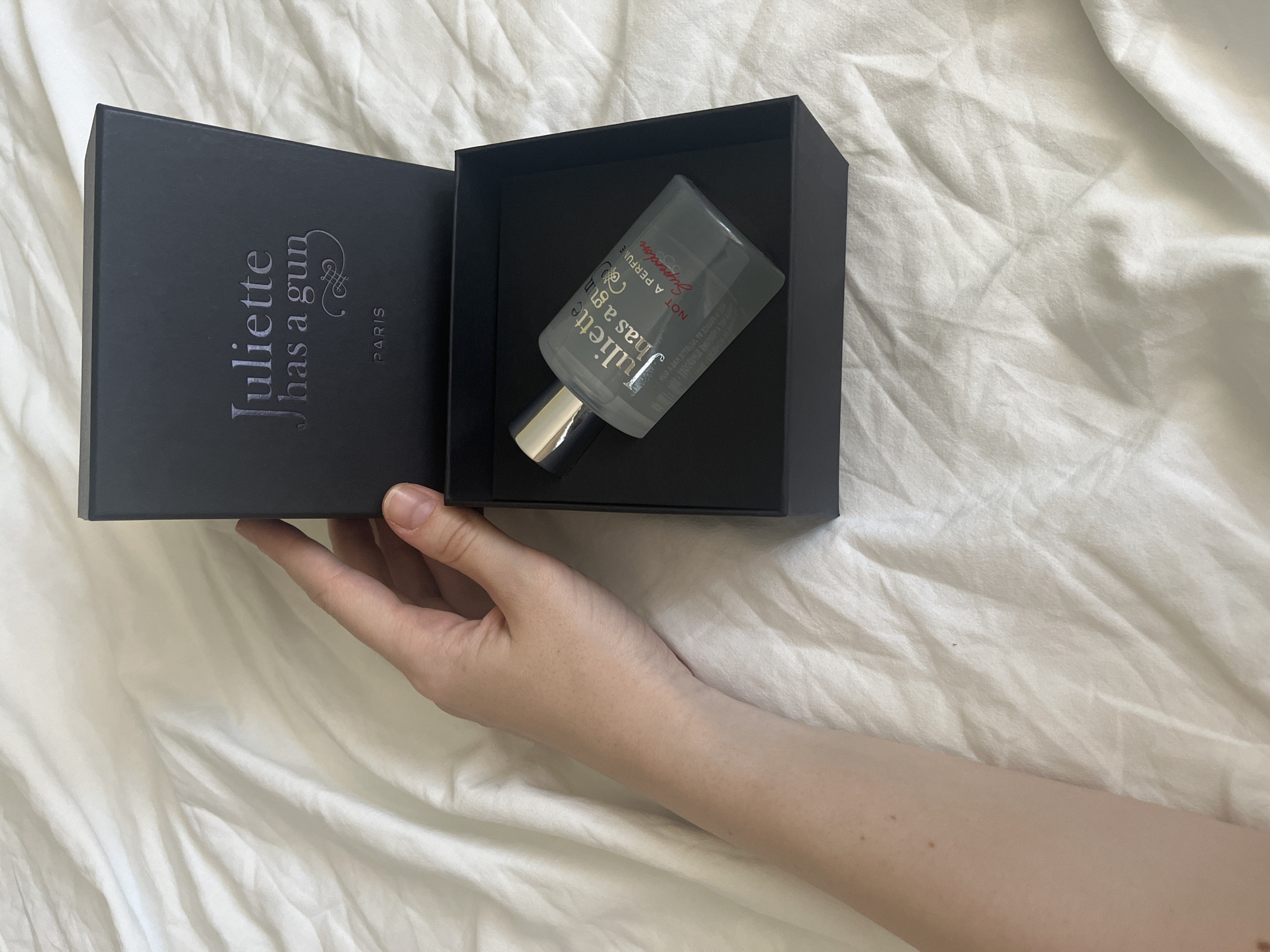 An overhead shot shows a hand holding open a black box, revealing a bottle of perfume inside. The box is embossed with the brand name "Juliette has a gun" and "PARIS". The perfume bottle is clear glass with a silver cap and a label that reads "Juliette has a gun NOT A PERFUME Superdose". The box is placed on a white, crumpled sheet. The image is taken from an eye-level perspective, with the hand entering the frame from the left. The lighting is soft and natural. There are no indications of the city of Komsomolskoye, Ukraine.
