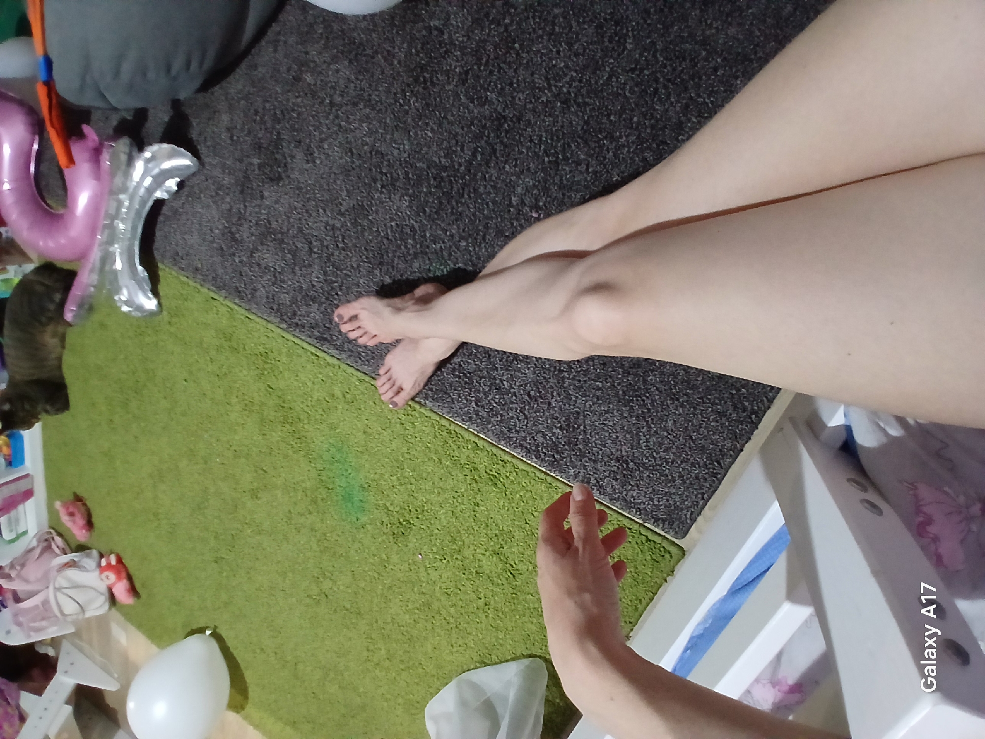 This indoor image captures a partial view of a person relaxing on a floor covered by two distinct rugs. The person's bare legs are prominent, crossed at the ankles, with dark nail polish visible on their toes. One hand is extended towards the lower right corner of the frame.
The setting appears to be a home in Yekaterinburg, Russia, featuring a dark gray, shaggy carpet covering a large area, bordered by a brighter green, shaggy rug. In the upper left, a gray cushion or pillow is visible next to what appears to be a partially deflated pink foil balloon, shaped like a letter (possibly 'U' or 'P'). Adjacent to it is a silver foil balloon, also appearing deflated, shaped like another letter (possibly 'Y' or 'V'). A striped cat is partially visible on the green rug, near the balloons.
In the lower part of the green rug, a white inflatable balloon rests. To the bottom right, a white piece of furniture, possibly a storage unit or shelf, holds various items, including pink fabrics and small pink stuffed toys. A blue and white patterned fabric or blanket is tucked underneath this unit. The lighting suggests an indoor environment, possibly with artificial light, given the shadows. The overall activity seems to be one of casual relaxation. The text "Galaxy A17" is visible in the bottom right corner of the image, indicating the camera used.