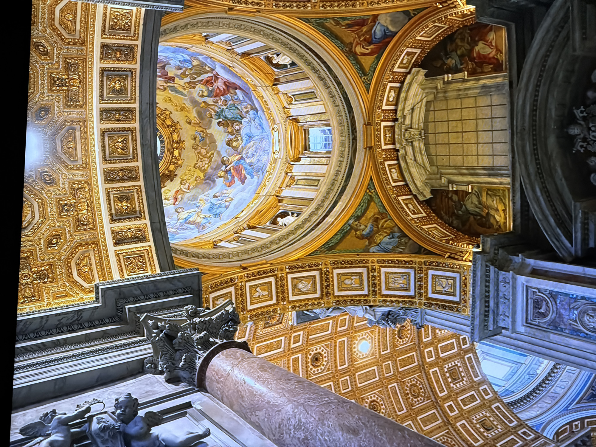 The image captures an ornate interior, likely a cathedral or basilica, characterized by its opulent decoration. A large, marble-patterned column dominates the left foreground, tapering upwards and partially obscuring the view of the surrounding architecture. Above and to the right, a gilded coffered ceiling stretches out, adorned with intricate patterns and smaller framed artworks.
The central focus is a magnificent dome, intricately painted with a fresco depicting a celestial scene. Numerous figures, including angels and human-like beings, are rendered in vibrant colors against a sky-blue backdrop, surrounded by golden accents. The dome is framed by golden drapery-like elements and pilasters.
Beneath the dome, a large, grid-patterned window is visible within an elaborate architectural setting. Flanking this window are painted murals, depicting figures in traditional attire. Further down, another arched architectural feature, also adorned with reliefs and a dark, textured surface, is present. Sculptural elements, possibly cherubs, are visible on a ledge in the upper left. The overall impression is one of grandeur, religious significance, and meticulous artistry. The scene is devoid of people and any indication of a specific time of day.
This image does not depict Chirchik, Uzbekistan.
