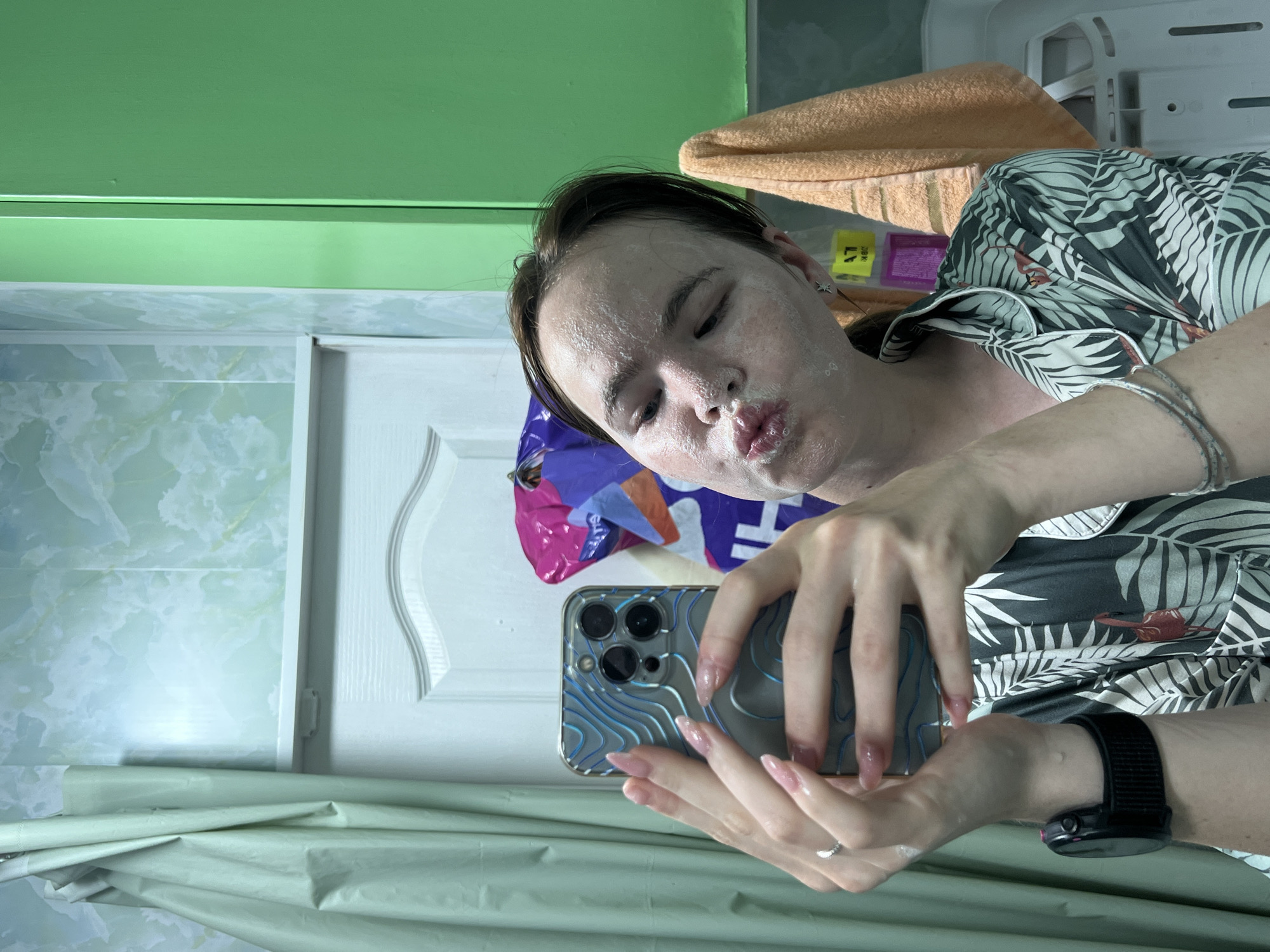 A young woman with fair skin and brown hair is taking a mirror selfie in a bathroom. She is wearing a dark green pajama top with a palm leaf print. Her face is covered in a white facial mask, and she is puckering her lips. She is holding a silver iPhone with a blue and silver patterned case in her hands. She is wearing a silver ring on her left ring finger and a silver bracelet on her left wrist. A black smartwatch is visible on her left wrist. The bathroom has light blue and green walls, a white door, and a light green shower curtain. An orange towel is hanging on the right side of the image. The location is not identifiable as Chaikovsky, Russia.