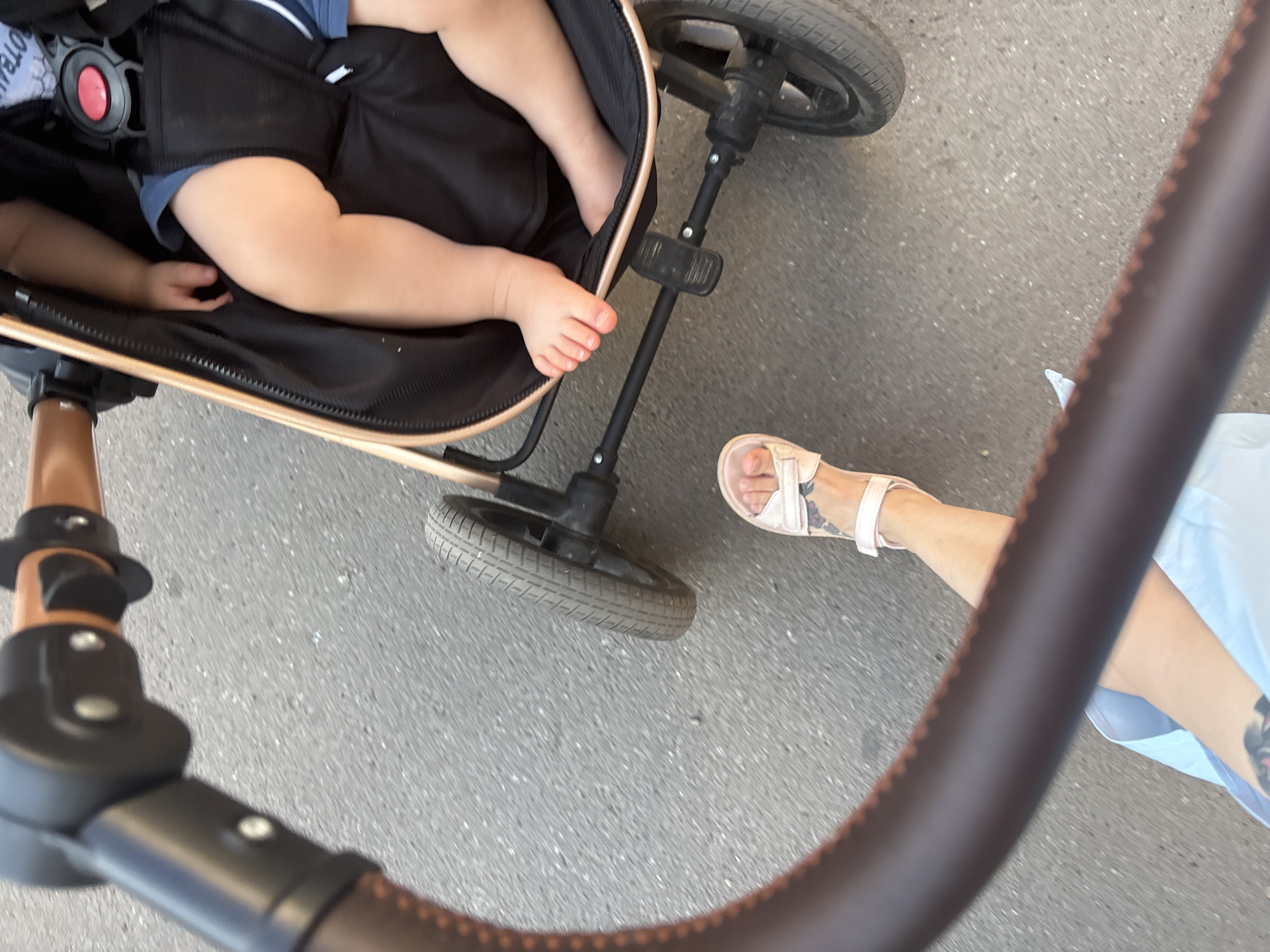 The image depicts a first-person perspective of a person pushing a stroller on a paved surface, likely a sidewalk or road, in Naberezhnye Chelny, Russia. The stroller's dark fabric interior is visible, with a child's legs and feet, wearing blue shorts, resting inside. The stroller features a brown leather-wrapped handle and a gold frame. A person's leg, wearing a light-colored sandal, is visible in the foreground, suggesting the photographer is the one pushing the stroller. The ground is asphalt with visible gravel.