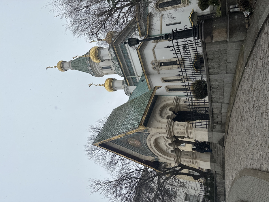 An exterior, low-angle, wide shot captures the St. Nicholas the Wonderworker Russian Church in Sofia, Bulgaria, on an overcast, snowy day. The church features a white facade, multiple green-tiled roofs, and five golden onion domes, each topped with a golden Orthodox cross. The main entrance, visible on the left, is an ornate arched portal with columns, topped by a gabled roof section featuring a mosaic in its tympanum. Fine snowflakes are visible against the muted sky and throughout the scene.
In the foreground, a curved cobblestone path slopes upwards towards the church. A dark grey stone retaining wall topped with a black ornamental metal fence separates the path from the elevated church grounds. Two individuals are partially visible at the base of the church's entrance steps, obscured by shadow and the architecture; one appears to be standing, clad in dark winter clothing, while the other is less distinct. Bare deciduous trees with dark branches frame the church on both sides. A black, ornate street lamp post stands prominently on the right, next to the fence and retaining wall. The overall atmosphere is cold and subdued.