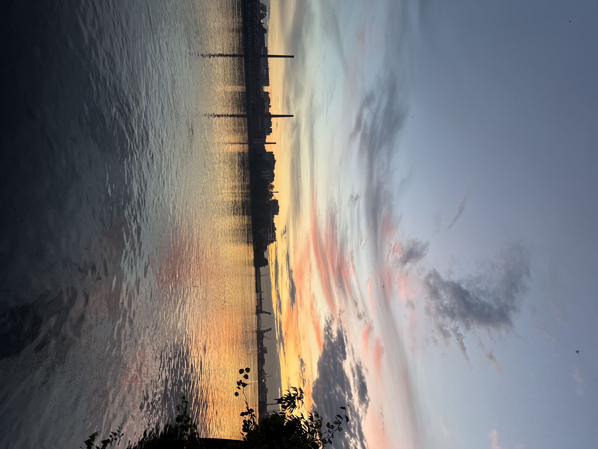 The image depicts a sunset over the Neva River in Saint Petersburg, Russia. Silhouetted against the colorful sky are industrial buildings and chimneys, suggesting an industrial district. The water reflects the warm hues of the sunset, creating a shimmering effect. In the distance, a bridge with raised bascules is visible, indicating it is open for navigation. The sky is a gradient of blues, grays, and oranges, with wispy clouds. A dark tree branch with leaves is visible in the foreground on the right side of the image. The overall scene is tranquil, capturing the end of the day in an urban riverside setting.