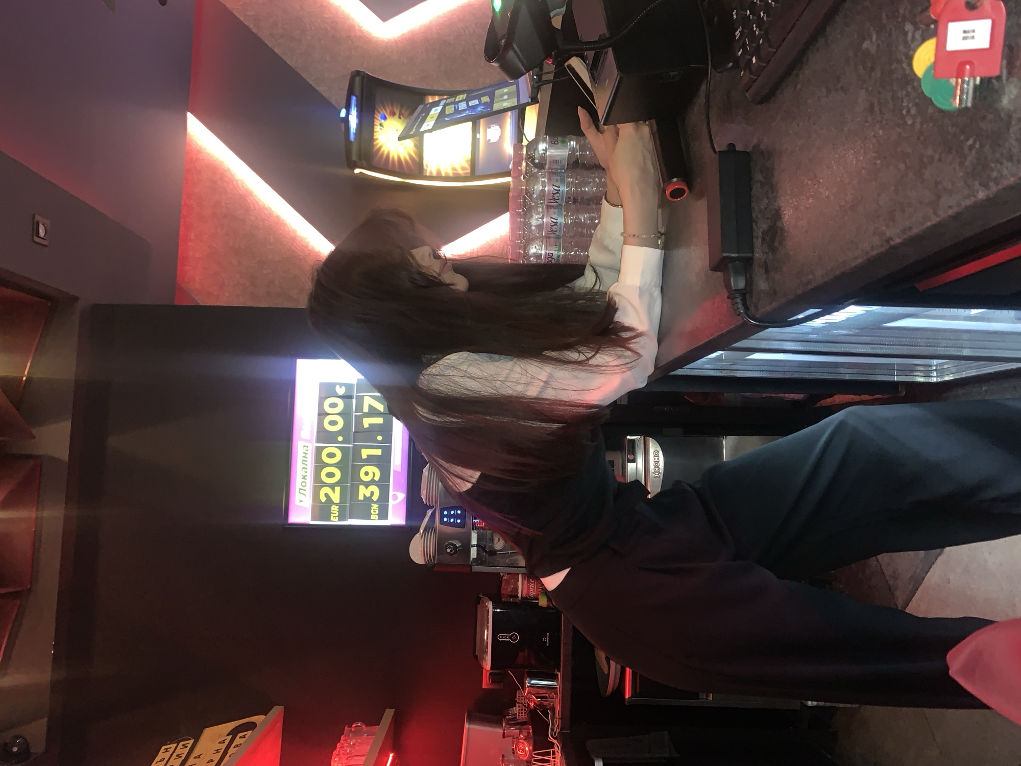 The image depicts a woman standing behind a counter in what appears to be a bar or entertainment establishment. She is wearing a dark vest over a white shirt and dark, wide-legged pants, leaning forward and resting her arms on the counter. In front of her, on the counter, is a cash register or payment terminal and several bottles of water. Behind her, a digital display shows currency exchange rates: "EUR 200.00" and "BGN 391.17". To the right, a gaming machine with a bright display is visible. The background features red and gray textured walls with neon lighting, typical of a modern entertainment venue. The overall atmosphere suggests a commercial setting, possibly a casino or a bar with gaming facilities in Burgas, Bulgaria.