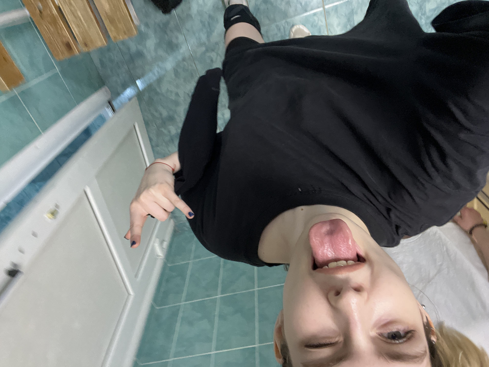 A selfie taken from a high angle shows a person sticking out their tongue and winking. The person is wearing a black long-sleeved shirt. The background includes green-tiled flooring, part of a white door, and what appears to be a wooden structure. It is not possible to confirm the location in the city of Vsevolozhsk, Russia, with this image alone.