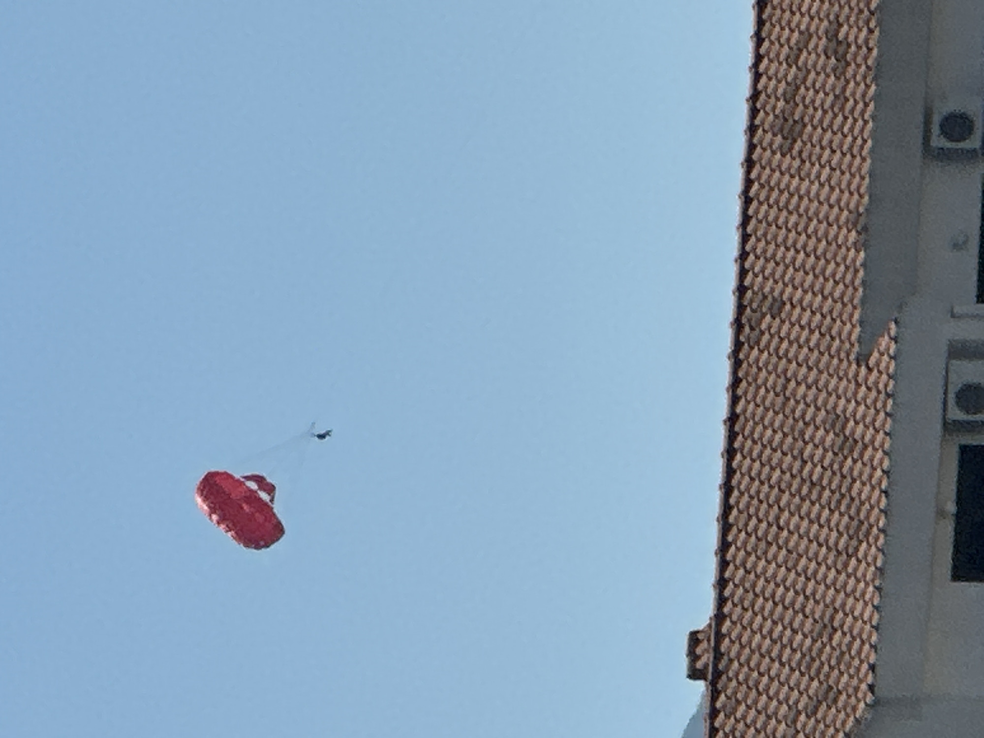 A person is parasailing against a clear blue sky, with a red parachute overhead. Below the sky, a tiled roof of a building is visible, and a mountain peak can be seen in the background. The scene suggests a leisure activity, likely in a coastal area given the presence of parasailing and a beach resort-like building.