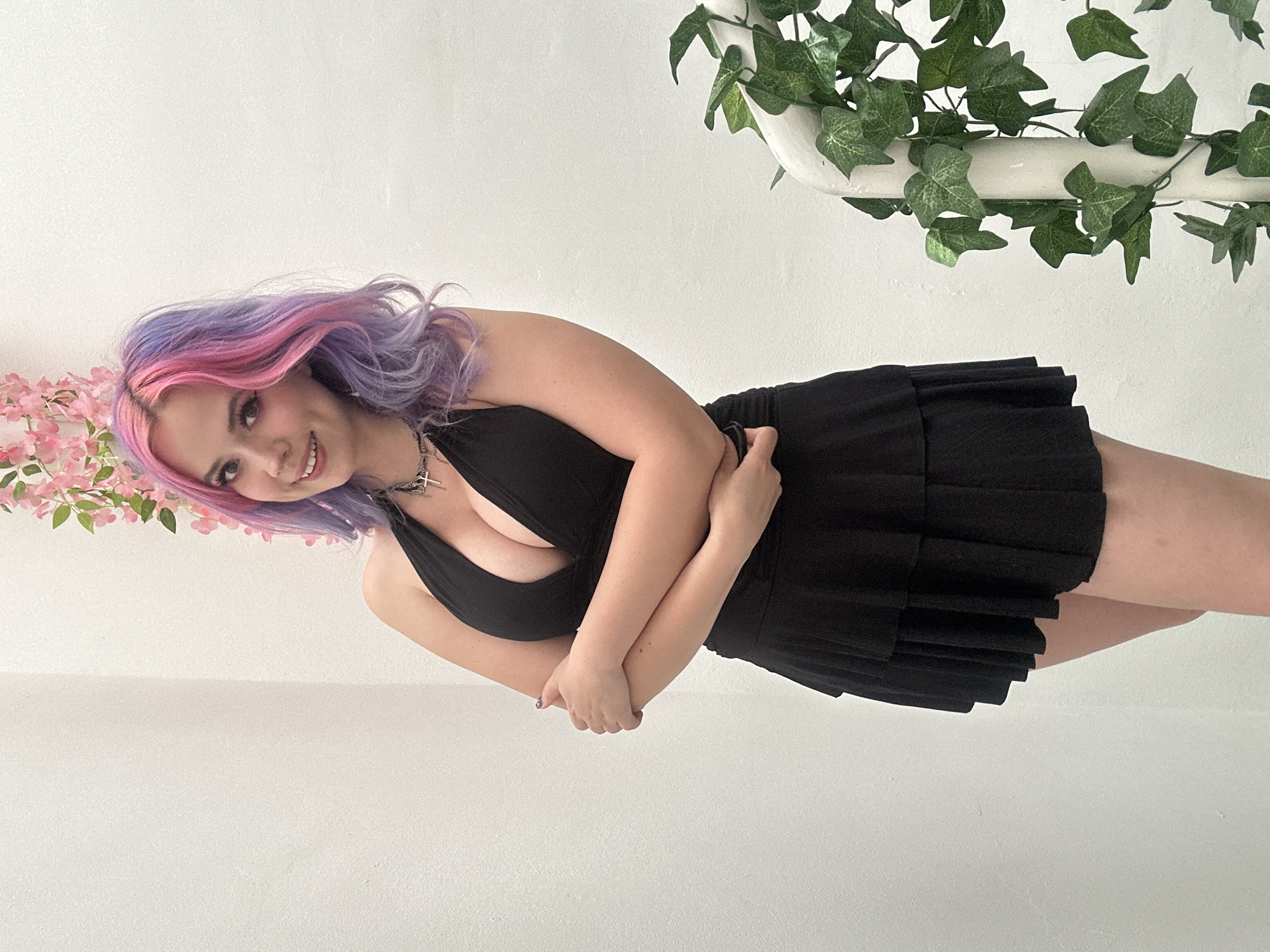 A young woman with long, wavy hair, dyed in vibrant shades of pink and purple, is the central subject of this brightly lit, indoor portrait. She appears to be in her late teens or early twenties, with a cheerful smile directed towards the camera. Her dark eyes are accentuated with makeup.
She is wearing a black halter-neck top or dress with a deep V-neckline, paired with a short, pleated black skirt. A silver-toned necklace with a cross pendant is visible around her neck. Her arms are crossed in front of her chest, with one hand gently holding the opposite forearm.
The background consists of a plain white wall, suggesting a clean, simple setting like a studio or an uncluttered room. Artificial pink floral branches, resembling cherry blossoms, are positioned to the left of her head, adding a soft, colorful element. In the upper right corner, green ivy-like vines are draped over a white pipe or rail, contributing a natural, albeit artificial, touch to the scene. The overall lighting is bright and even, casting minimal shadows.
The woman's pose and expression convey a sense of confidence and friendliness. There is no visible text in the image. While the location context is given as Zagreb, Croatia, there are no specific visual cues within the image itself to confirm this.