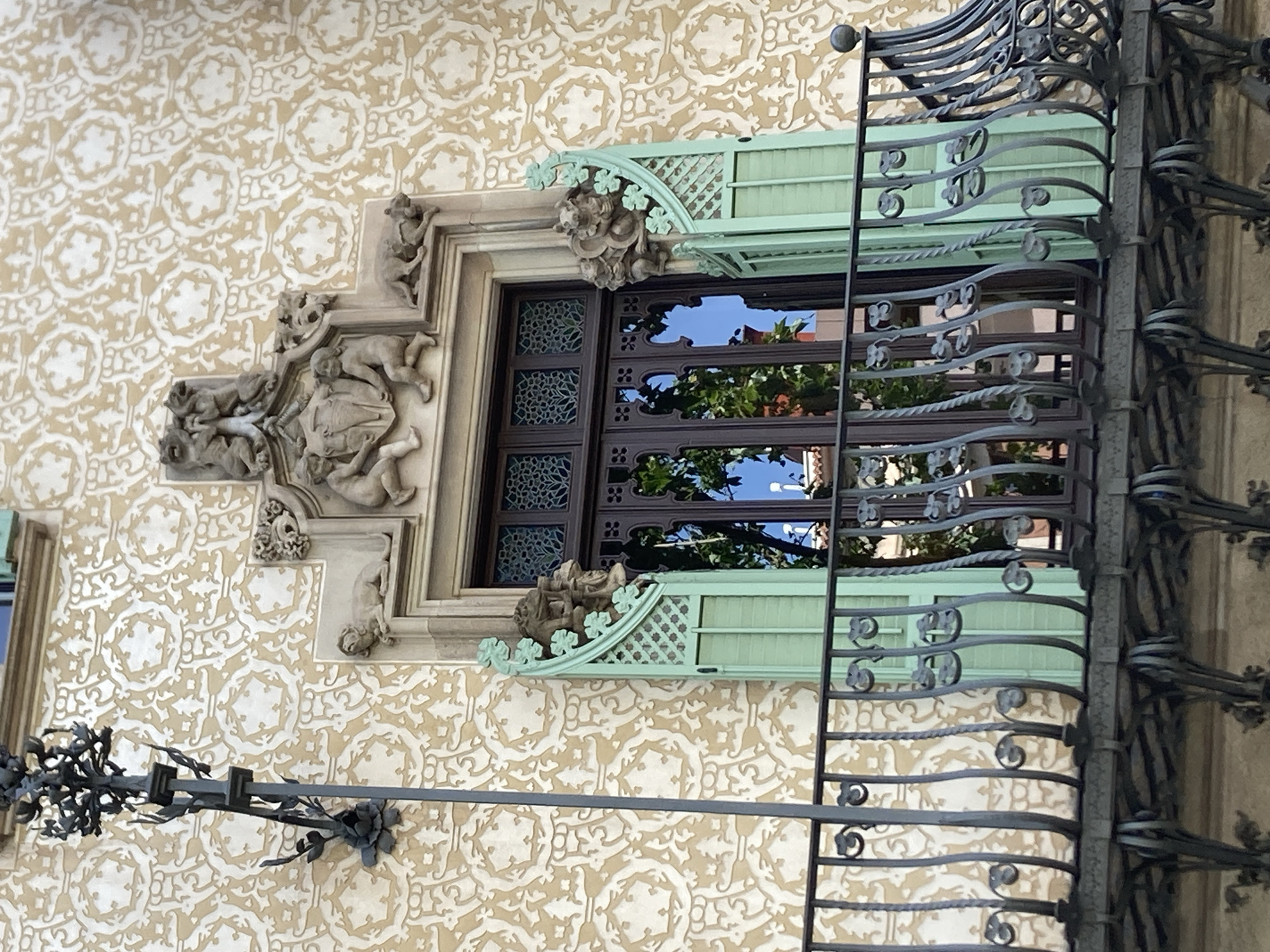 This is a close-up, eye-level outdoor shot of a window on a building in Barcelona, Spain. The window itself is intricately designed with a dark brown frame and stained glass panes featuring a repeating floral pattern. It is framed by ornate stone carvings on the left and a light green decorative metal structure on the top. A dark metal railing with elaborate scrolled details runs vertically in front of the window, partially obscuring it and extending to the right of the frame.
The building's facade is covered in a textured wallpaper or plaster with a repeating pattern of stylized floral or star-like motifs in a light tan color against a cream background. The stone carvings around the window depict cherubic figures and what appear to be animals or mythological creatures, suggesting an Art Nouveau or similar architectural style. The green metalwork above the window is also decorative, with a lattice-like design and clover-shaped embellishments.
The reflection in the window glass shows greenery from trees and a bright blue sky, indicating it is daytime with clear weather. The overall impression is one of architectural detail and artistry. No text is visible in the image.