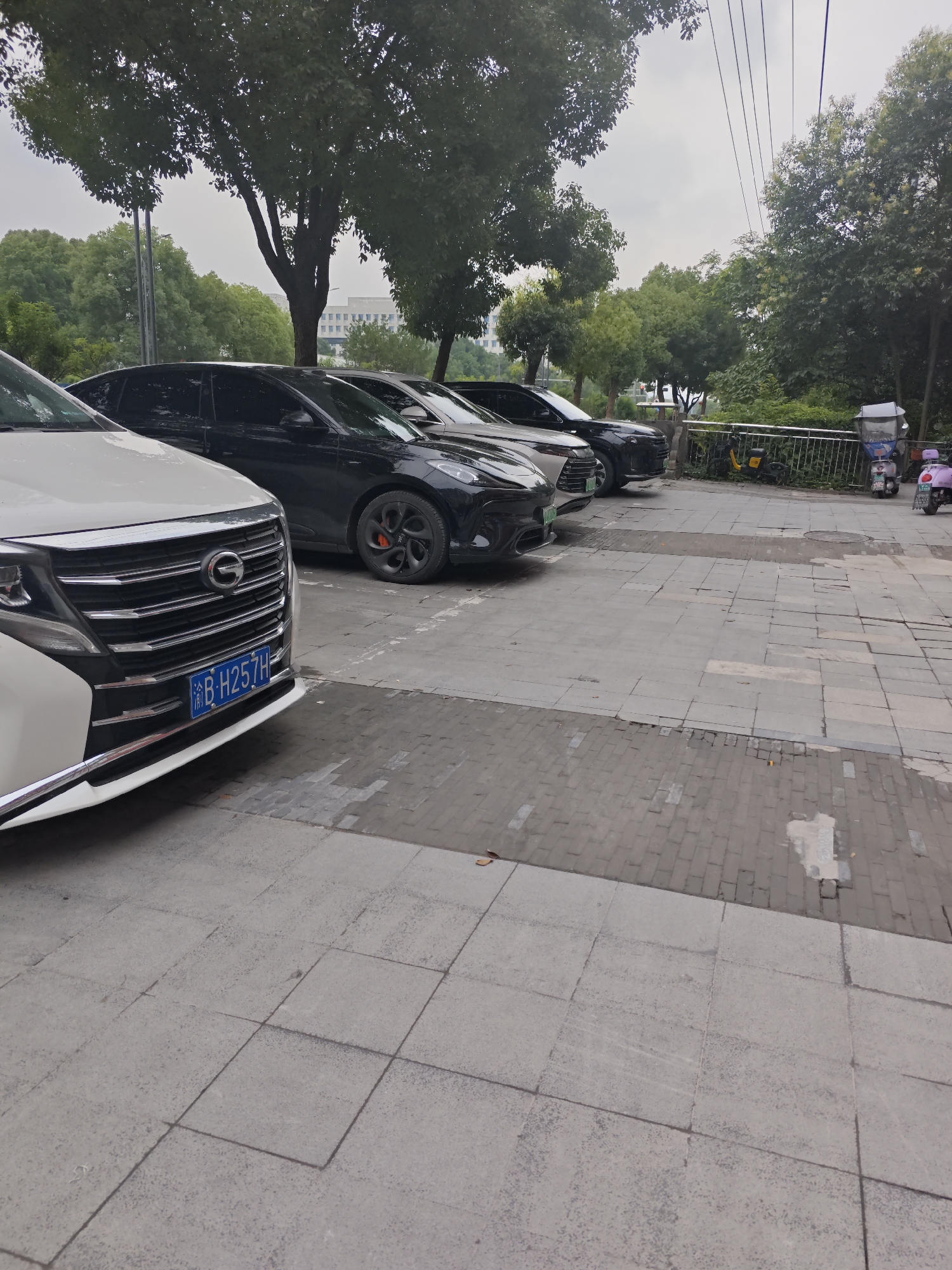 A white SUV is parked in the foreground on a paved area. Behind it, a line of black sedans is also parked. In the background, there is a row of trees, a metal fence, and three parked electric scooters. The sky is overcast. The license plate of the white SUV reads "渝B H257H". The location is the Chongqing High-tech Zone, China.