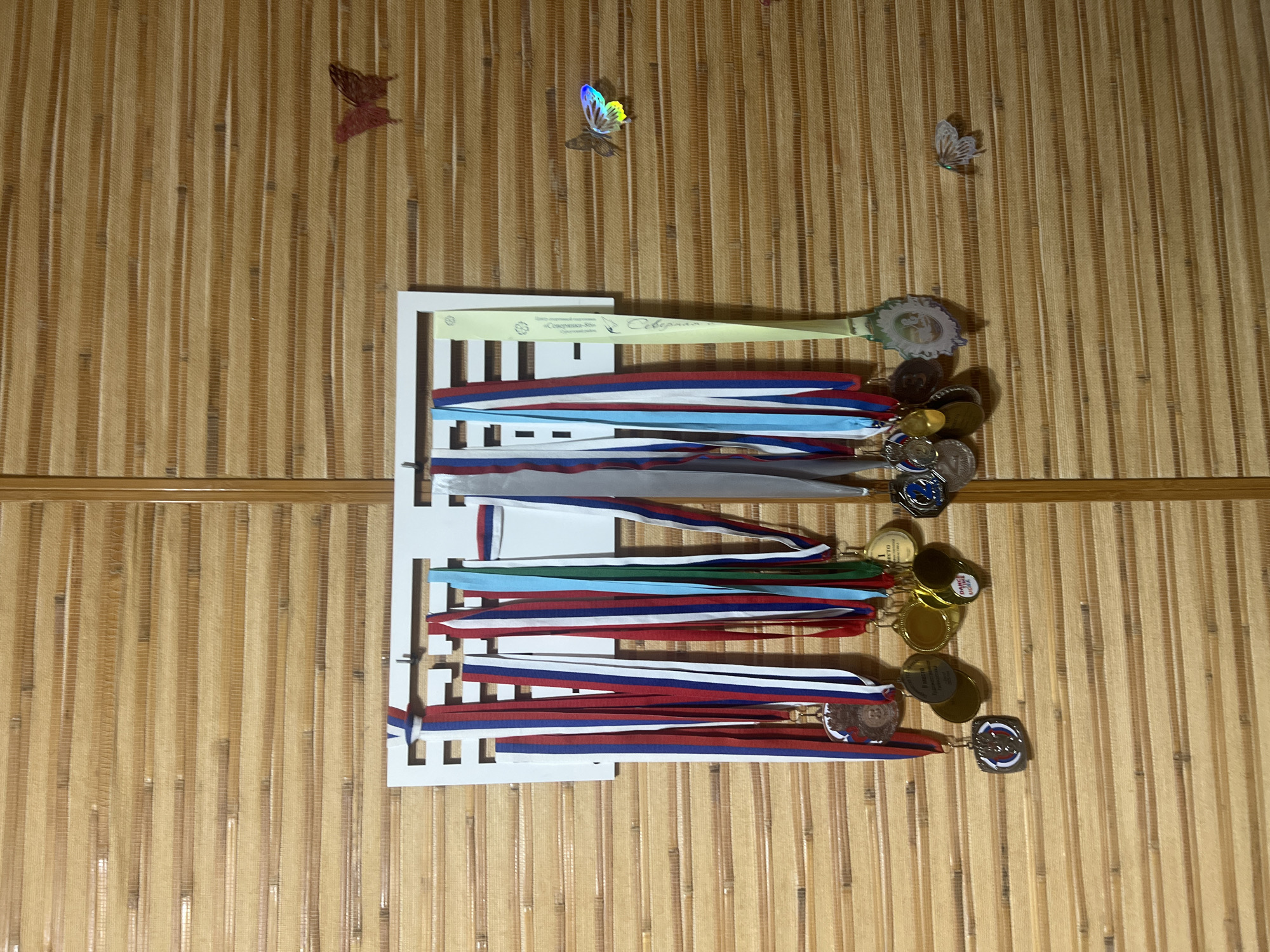 The image shows a wall decorated with a horizontal bamboo-like textured covering. A white rack is mounted on the wall, displaying a collection of medals with various colored ribbons, predominantly in red, white, and blue. There are also decorative butterflies adhered to the wall. The scene appears to be indoors, possibly in a residential setting, and given the prominent Russian flag colors on the ribbons, it could be located in Surgut, Russia. No individuals are present in the image, and no actions are being performed. The lighting suggests it is daytime.