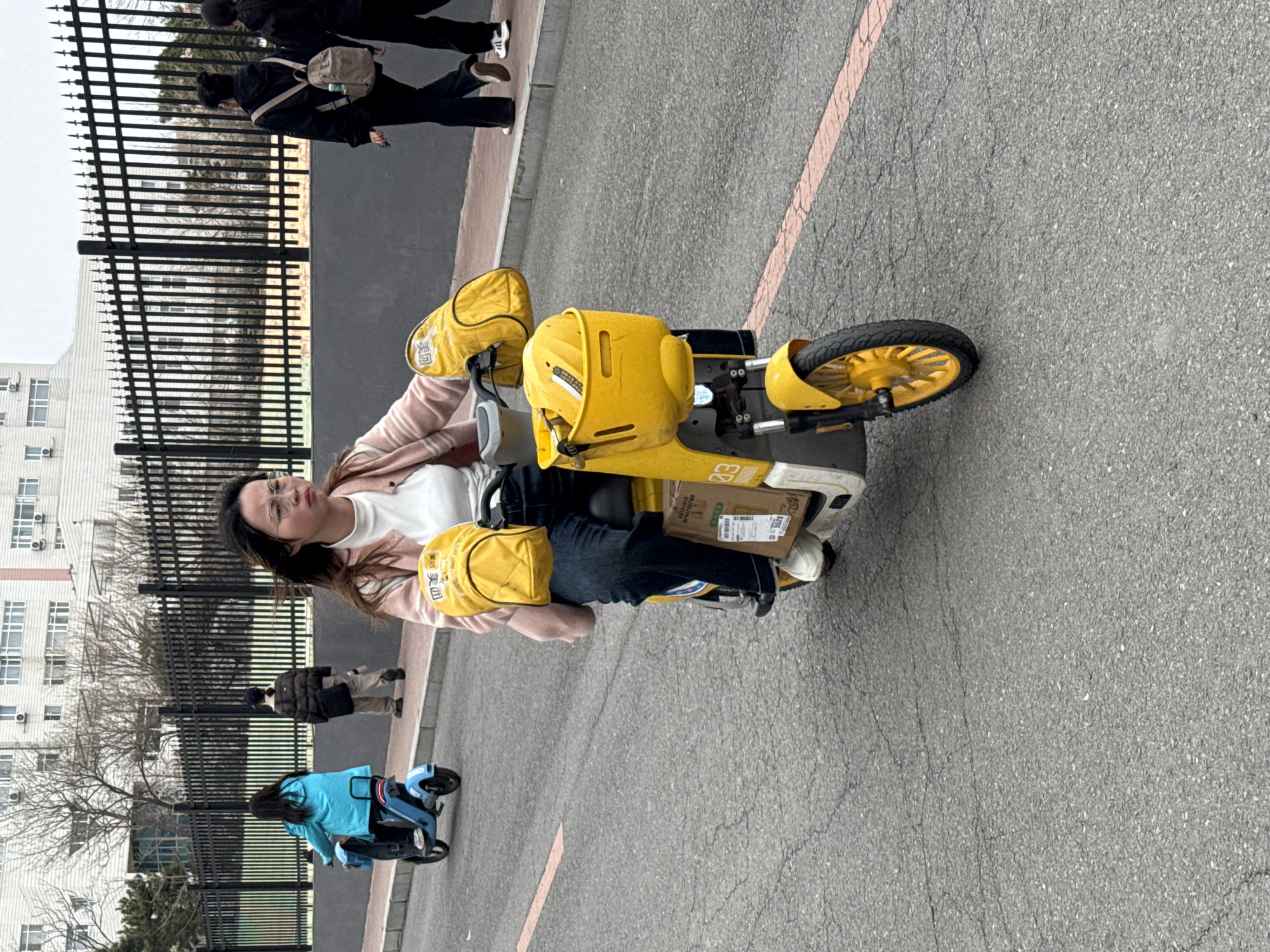 A woman is sitting on a yellow electric bicycle in Zhongtun, China. She wears glasses, a white t-shirt, pink sweater, and jeans. Two yellow bags with Chinese writing are attached to the front of the bicycle. There's a cardboard box on the rear rack. A blue electric bicycle is parked nearby. A black metal fence is to the left with a building visible behind it. Several pedestrians are on the sidewalk. The road is asphalt with a red line marking a lane.