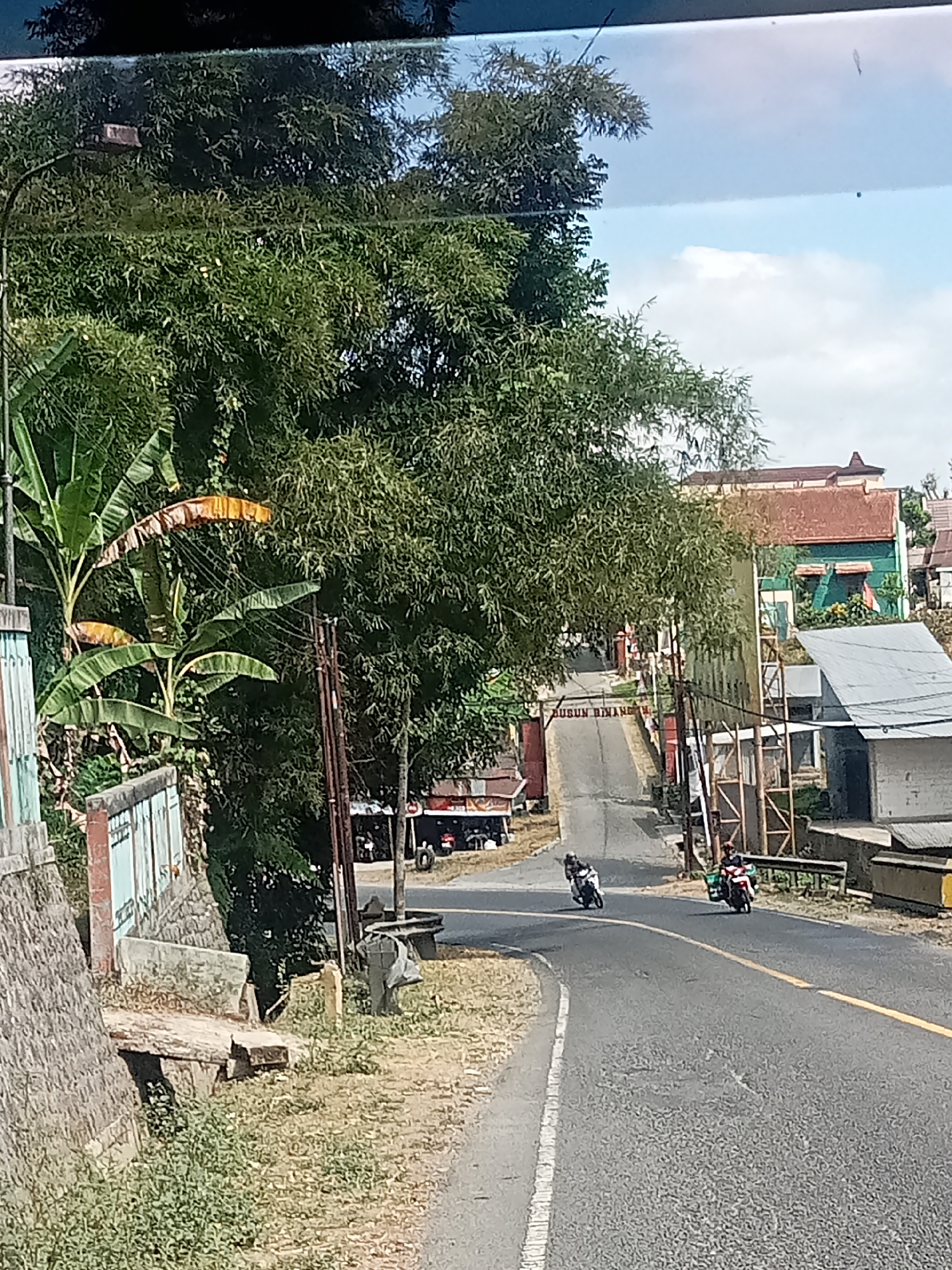 The image captures a street scene in Selomerto, Indonesia. A paved road curves through a landscape dominated by lush green trees and vegetation. In the foreground, a stone wall and some vegetation line the left side of the road. Further up the road, there's an arched gateway with text that appears to read "Dusun Binanga". On the right side of the road, various buildings and structures are visible, including houses and what looks like a small shop. Two motorcycles are traveling on the road; one in the foreground carrying a single rider, and another further up the road with two riders. The sky is partly cloudy, suggesting daylight. The overall impression is of a rural or semi-rural setting within the town.