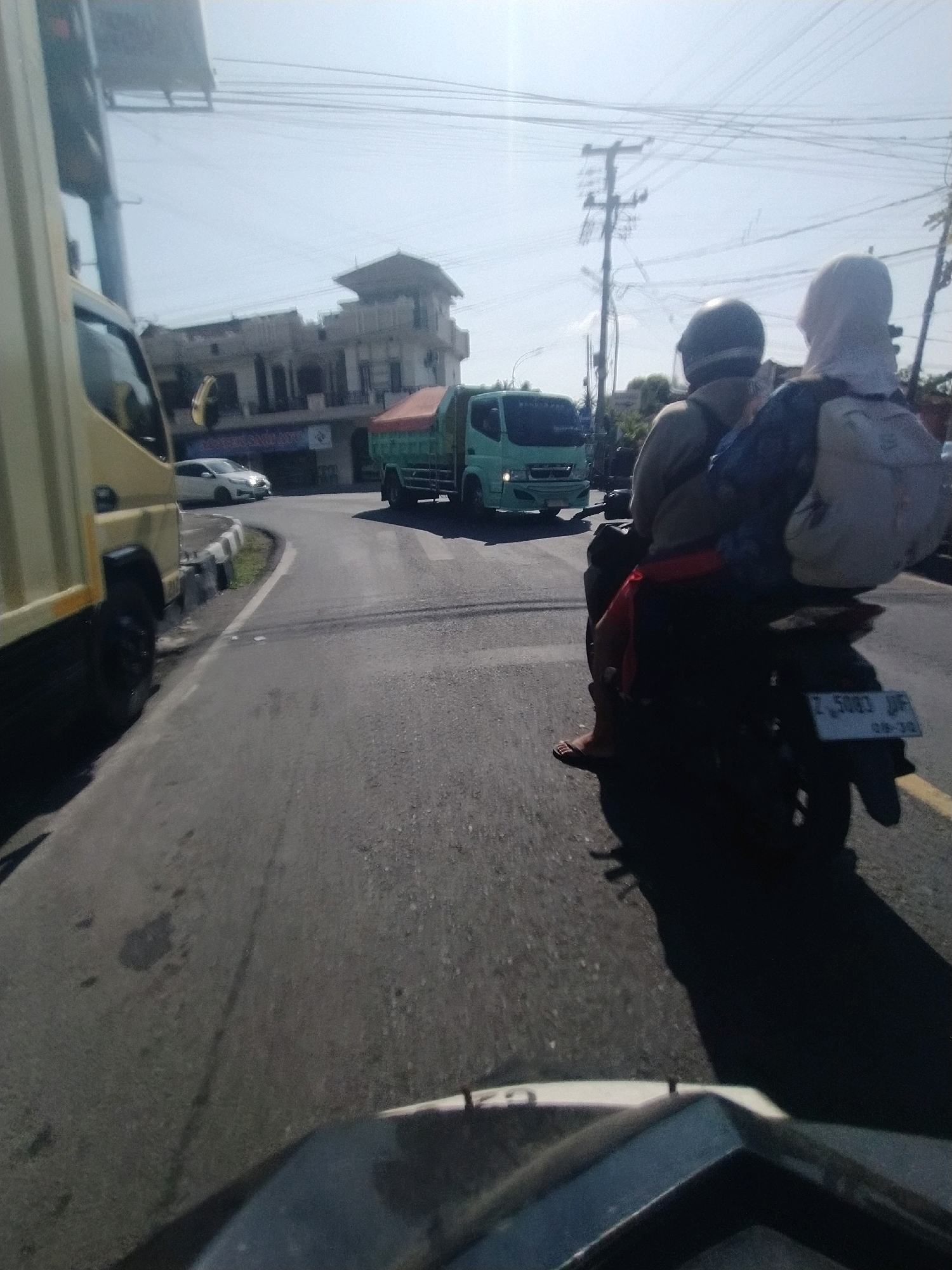 A side view of a sunny road in what appears to be a rural or semi-urban area in Desa Karangbenda, Indonesia, shows a light-colored truck in the foreground on the left, obscuring some of the road and scenery. Further down the road, a light green truck is visible, with a red tarp covering its load. In the middle ground, a motorcycle carrying two people is traveling away from the camera. The person in the back, wearing a headscarf and backpack, faces away from the camera. The person in front, wearing a helmet, is also facing away. Their legs are visible, with one person wearing sandals. The road has visible lane markings, and utility poles with numerous wires are present along the side. Buildings with tiled roofs are in the background, and vegetation is sparse. The sky is clear and bright blue.