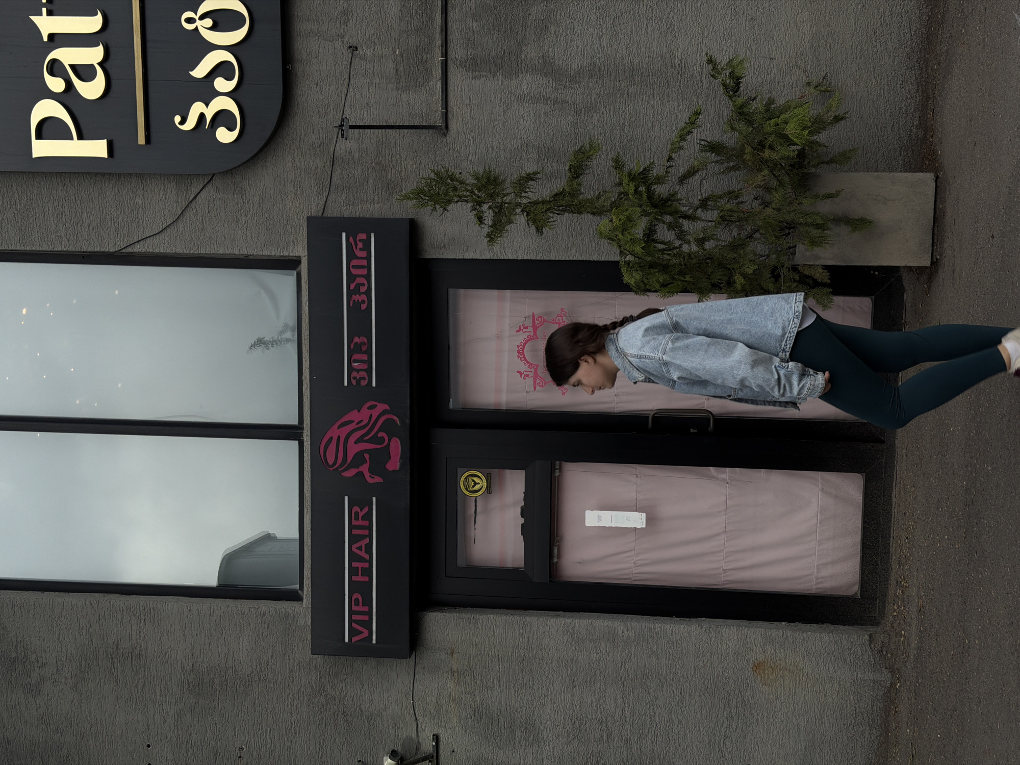 The image shows the exterior of a business, likely a hair salon called "VIP HAIR" located in Tbilisi, Georgia, as indicated by the Georgian script on the signage. The business has a modern sign with English and Georgian text, as well as an image of a stylized lion or griffin. To the left of the entrance is a large window. A young woman is standing in front of the entrance wearing a denim jacket and dark leggings. A small evergreen tree is planted in a concrete container next to the entrance. The building has a textured gray facade, and the sidewalk is visible on the right side of the frame.