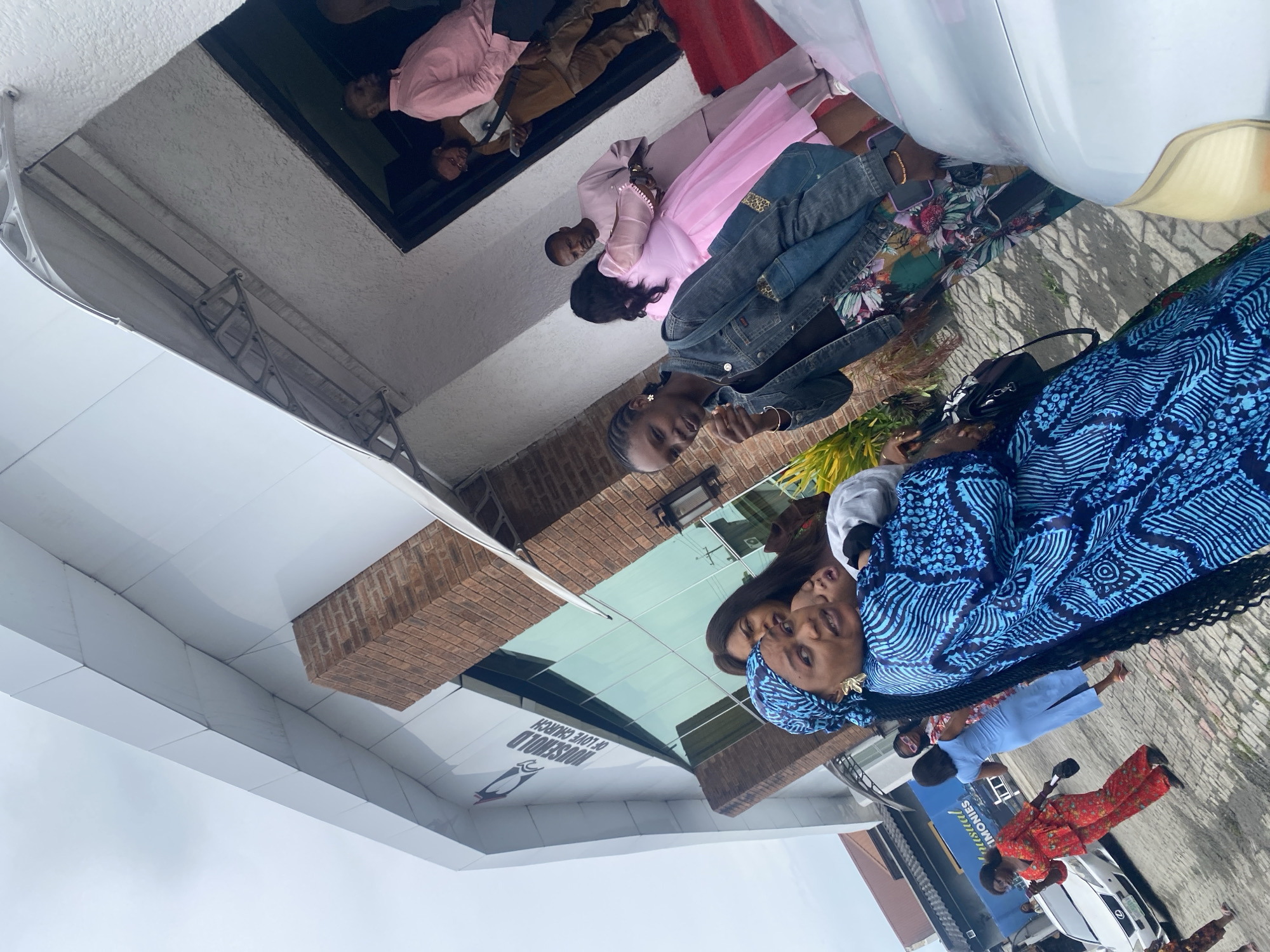 The image shows several people in front of a building in Ado, Nigeria. The building is labeled "Household of Love Church." A woman in a blue patterned outfit is prominent in the foreground, standing with another woman. Additional people are visible, including some in pink attire. A white car is parked nearby. The sky is overcast.