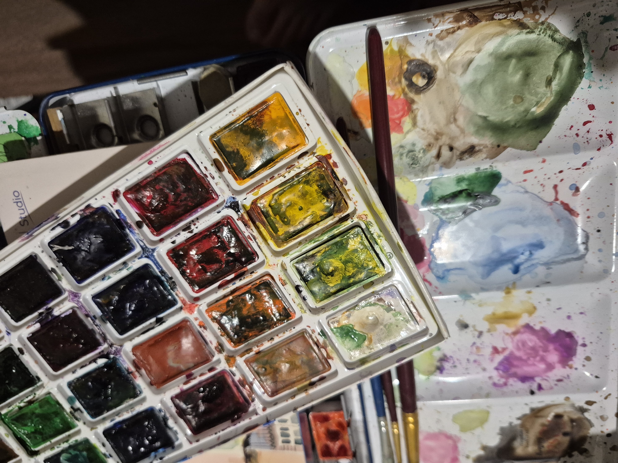 The image shows an overhead view of art supplies, including a watercolor paint set and a palette with mixed paints. The watercolor paint set is a rectangular white plastic case with 24 wells, many of which contain dried or partially used cakes of watercolor paint in various colors, including green, blue, purple, black, red, orange, brown, and yellow. Some wells show remnants of wet paint. A portion of a cream-colored booklet with the word "Studio" printed on it in blue lettering is visible at the top.
Below the watercolor set is a white plastic palette with several wells and a larger flat mixing area. This palette contains various pools of mixed watercolor paints in shades of pink, purple, blue, green, brown, and yellow. There are also spatters of paint across the palette.
Several paintbrushes with wooden handles are resting on the palette and extending out of the frame. One brush has a dark red handle, and another has a yellow handle. The overall setting appears to be an artist's workspace, likely indoors.
The image does not contain any information that would allow for the identification of a specific location in Popovo, Bulgaria. There are no landmarks, street signs, or any other contextual clues that would place the scene in that city. The scene is entirely focused on the art supplies.