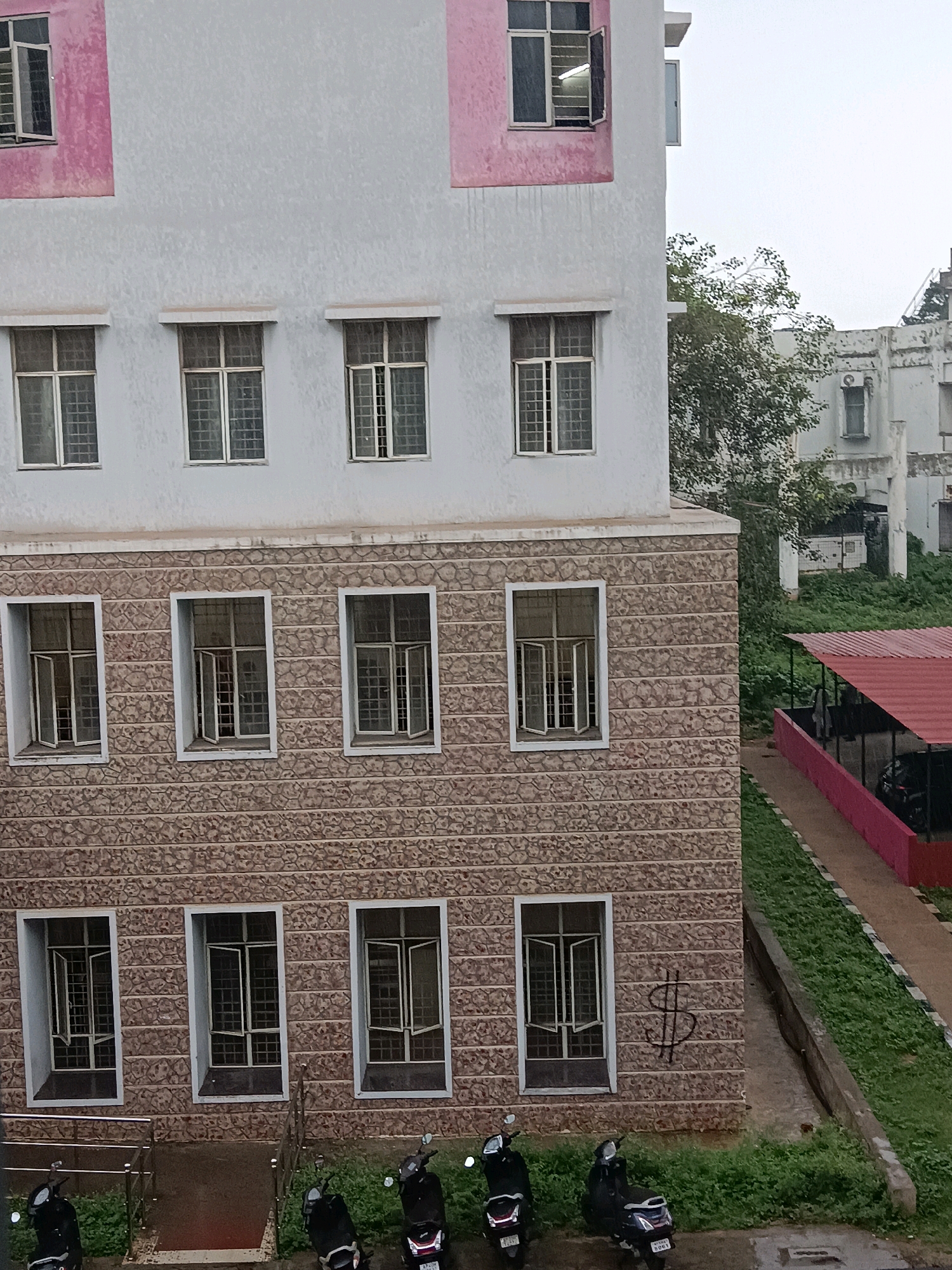 The image shows a multi-story building with a textured stone facade on the lower levels and a lighter colored facade with pink accents on the upper levels. The building has numerous windows, some of which are open. The lower part of the image shows several parked motorcycles on a paved area surrounded by grass. In the background, there is another building and lush greenery. The overall scene suggests an outdoor urban environment. The presence of the motorcycles and the style of the building are consistent with locations in Visakhapatnam, India.