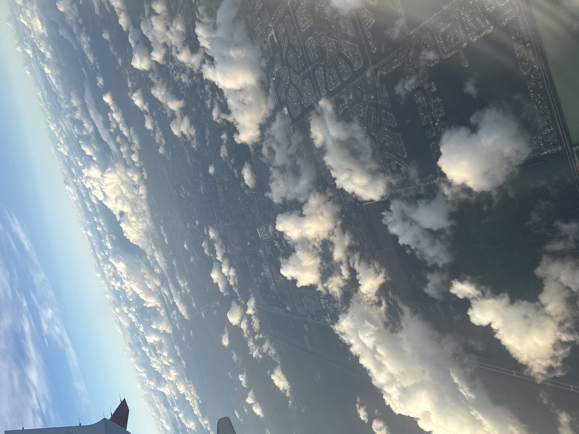 The image presents an aerial view, likely captured from an airplane. Below, a cityscape sprawls, partially obscured by a layer of cumulus clouds. The urban landscape shows a dense network of roads and residential areas, interspersed with patches of greenery. A highway is clearly visible cutting across the scene. The clouds vary in size and density, with some appearing thick and opaque, while others are more scattered and translucent, allowing glimpses of the ground below. In the foreground, parts of the aircraft, including a wing and the tail with a red emblem, are visible, indicating the perspective is from inside the plane. The sky transitions from a clear blue on the left to a hazy, sun-lit area on the right, suggesting early morning or late afternoon lighting. The location in the city of Mexico City, Mexico cannot be determined.