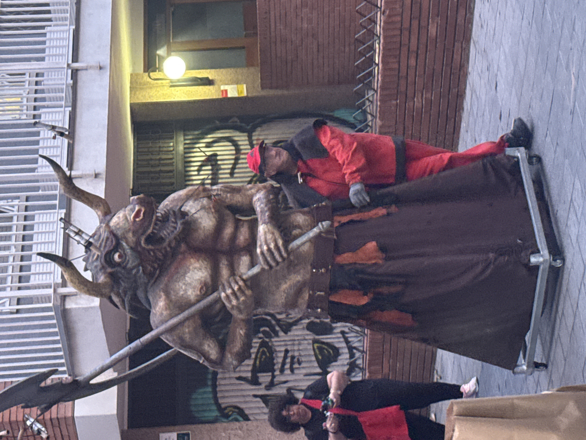The image displays an outdoor street scene in Barcelona, Spain, featuring a large, mobile, anthropomorphic figure with a bull-like head, muscular torso, and humanoid body. This central figure has prominent horns, glowing red eyes, sharp teeth, and holds a dark scythe-like implement in both hands across its chest. Its lower body is covered by a dark brown fabric skirt with distressed orange-brown accents, secured by a dark belt. The entire figure is mounted on a silver rectangular platform equipped with multiple small wheels.
To the right of the figure, a man is positioned, wearing a red baseball cap, a red jacket with a black cape-like overlay on the shoulders, dark gloves, black trousers, and black shoes. He appears to be interacting with or guiding the wheeled figure with his left hand near its side, looking forward and slightly left.
To the left and slightly behind the central figure, a woman with dark, curly hair is visible. She wears a black top, black trousers, and light-colored sneakers, holding small, colorful items in her hands. A red cross-body bag hangs from her shoulder, and she is looking towards the large figure.
The background consists of architectural elements including a light-colored building with a dark metal gate and a large, stylized black and green graffiti mural on an adjacent wall to the left. To the right, another building shows white-railed balconies on an upper floor, a spherical light fixture on a lower wall, and a section of a red brick wall with a low black metal fence in front of it. The ground surface is paved with grey interlocking tiles.
