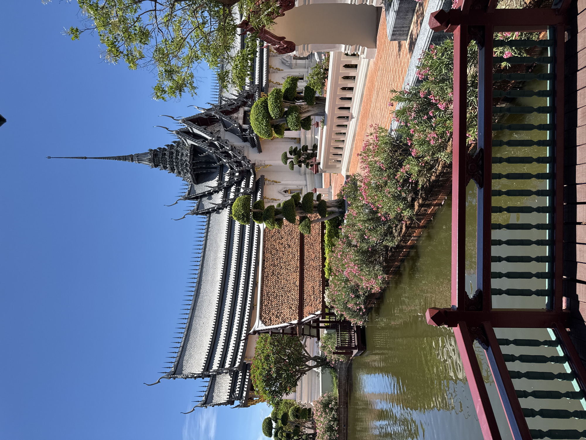 The image depicts Sanphet Prasat Palace, located in the Ancient City (Muang Boran) of Bang Pu Subdistrict Municipality, Thailand. The palace is characterized by its traditional Thai architectural style, featuring multi-tiered roofs with ornate detailing and a central spire reaching towards a clear blue sky. The palace is set against a backdrop of manicured landscaping, including decorative trees, flowering bushes with pink blossoms, and a reflecting pond. A portion of a wooden bridge or walkway with a decorative railing is visible in the foreground.