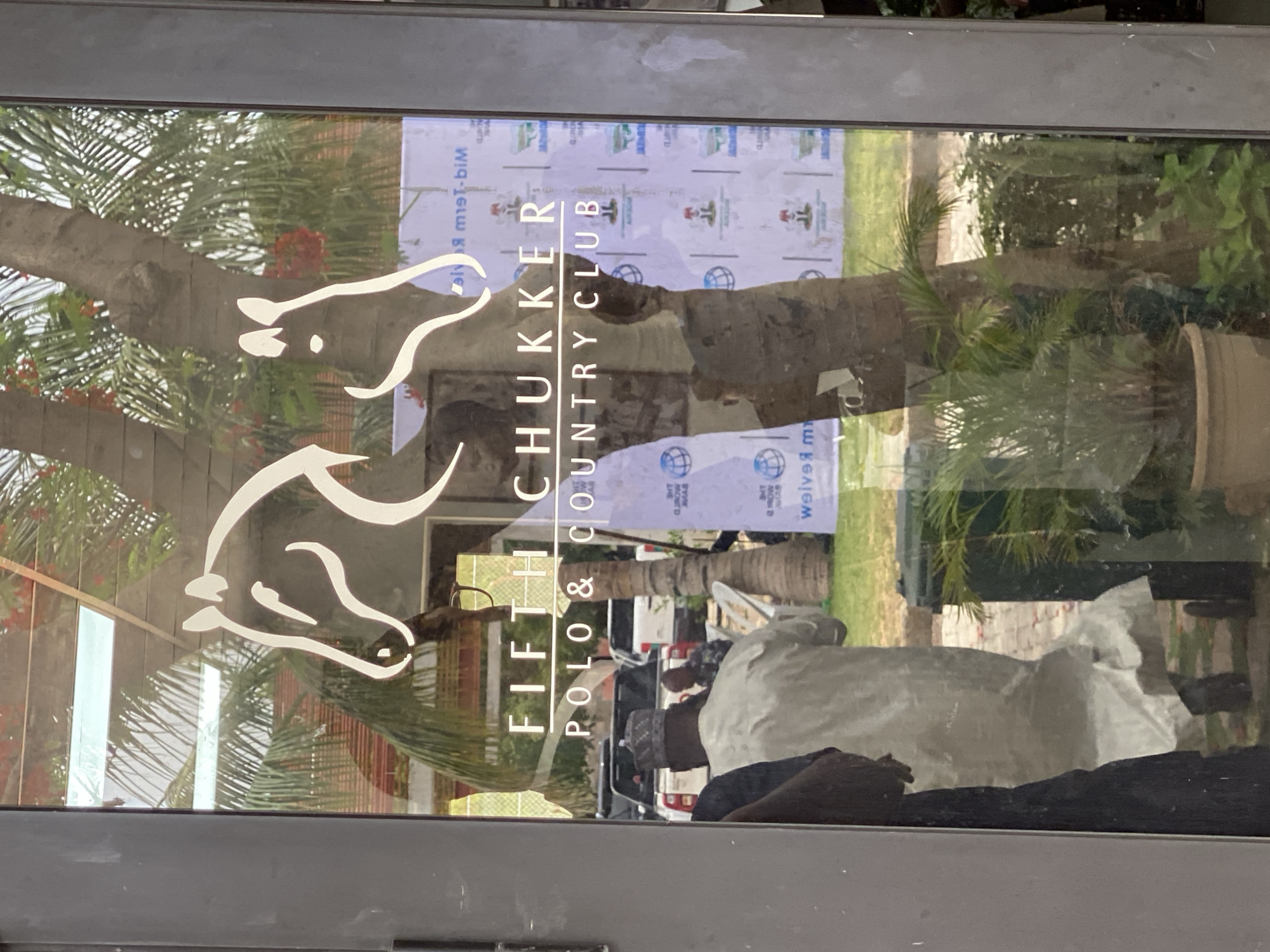 The image is a photo taken through a glass door or window, displaying prominent reflections of an outdoor scene and people, overlaid with branding. A white, stylized logo of two mirrored horse heads, one facing left and one right, is applied to the glass. Below it, the vertical text "FIFTH CHUKKER POLO & COUNTRY CLUB" is also visible in white.
The reflections indicate a bright, sunny daytime environment. Lush greenery, including palm trees and a tree with reddish-orange flowers, is prominent. In the background, a large white banner with repeated logos, such as a red cap/hat icon and a globe icon, and mirrored text (partially legible as "M-BIM" and "weive"), suggests an ongoing event or sponsorship. A grassy area, likely a polo field, and several parked vehicles are also reflected.
Multiple people are visible in the reflections. A person in a light-colored, possibly traditional, garment stands in the lower-middle, with another individual wearing a dark, patterned cap behind them. The scene depicts activity outside the entrance of the Fifth Chukker Polo & Country Club, located in Kwarau, Nigeria.