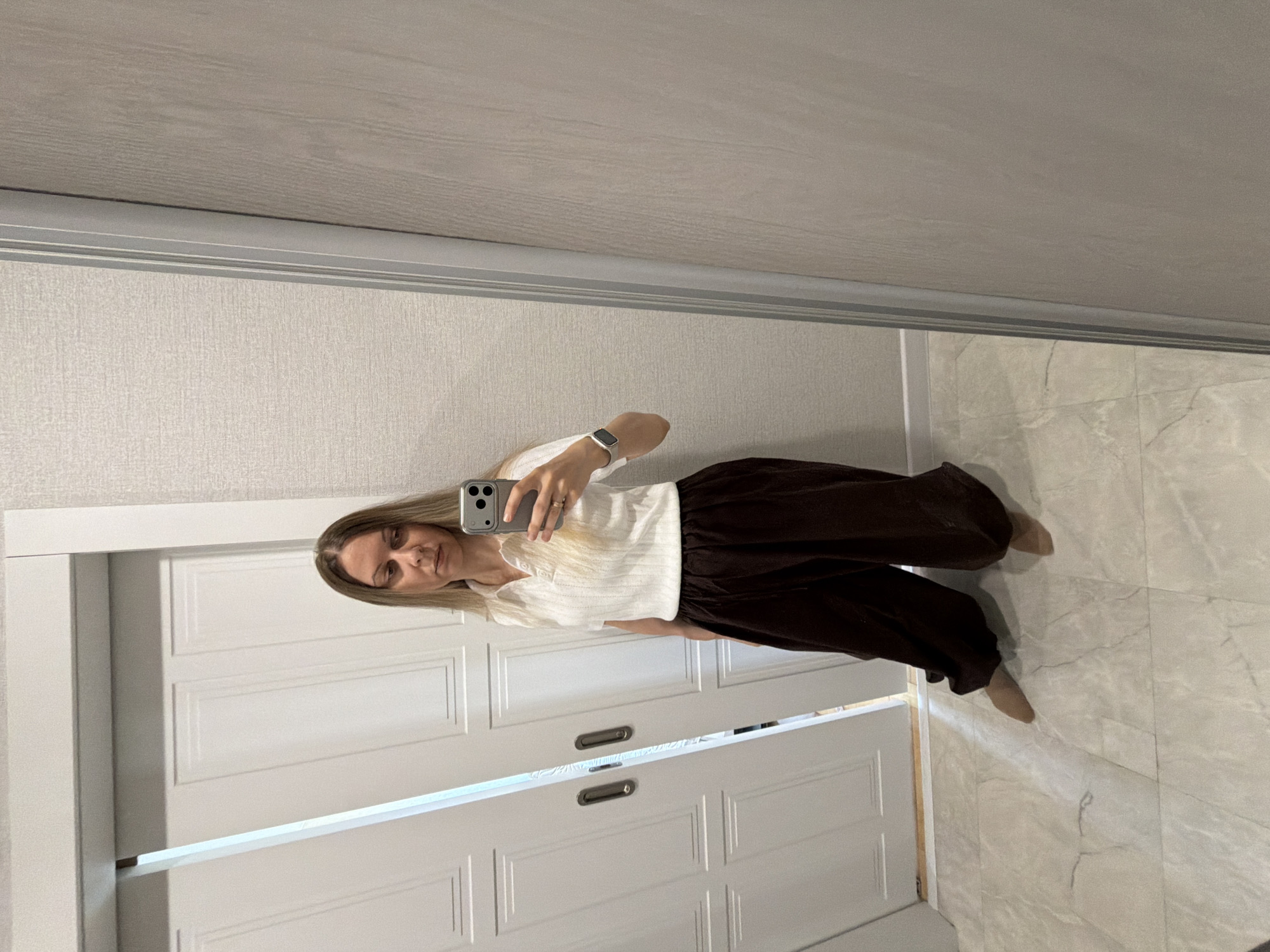 A woman with blonde hair is standing in what appears to be a residential hallway and taking a selfie in a mirror. She is wearing a white, ribbed polo shirt and dark brown, wide-leg pants. She has a watch on her left wrist. The hallway has light-colored, tiled flooring and white sliding doors in the background. The walls have a textured wallpaper. The scene is indoors, likely in a home. The specific location in Izhevsk, Russia cannot be determined from the image alone.