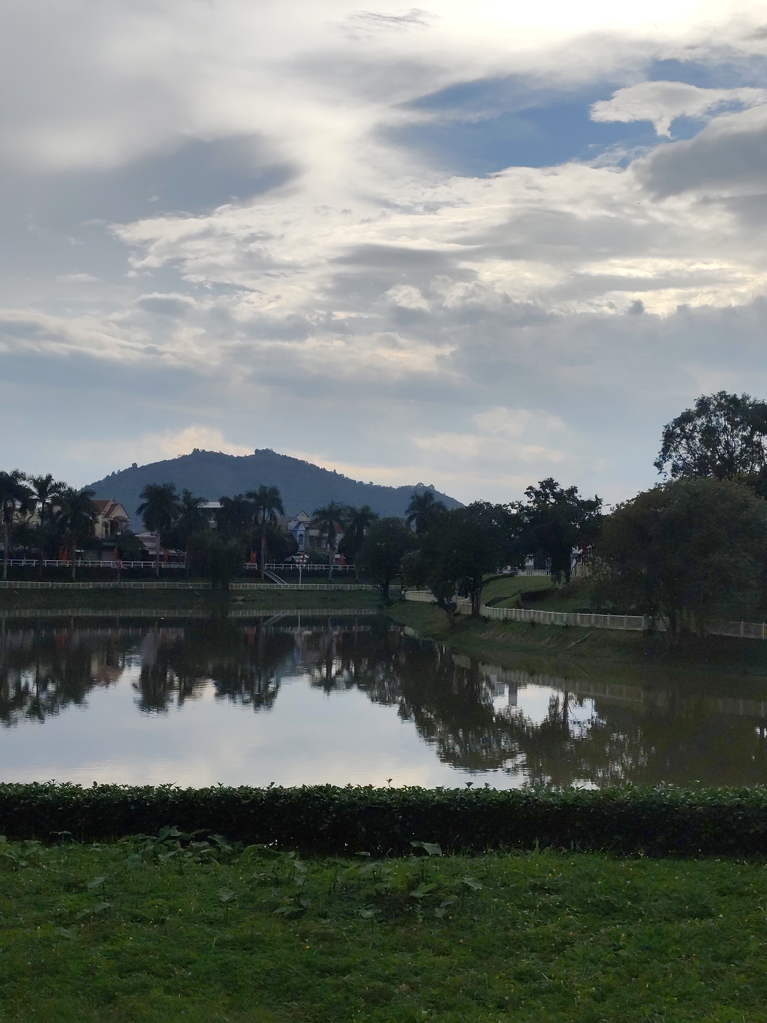 The image captures a serene landscape in Ma Đa Guôi, Vietnam, characterized by a tranquil lake reflecting the sky and surrounding scenery. The sky above is filled with dramatic clouds, ranging from light gray to darker shades, with patches of blue peeking through. The reflection on the calm surface of the lake mirrors the overcast sky and the silhouettes of trees and buildings on the opposite bank.
The mid-ground features a well-maintained bank along the lake, adorned with a white fence and lush green grass. Beyond the fence, a line of trees, including palm trees and other foliage, creates a natural barrier. Behind the trees, several houses with distinct architectural styles, some with red-tiled roofs and light-colored facades, are visible. In the distance, a prominent, rounded mountain covered in vegetation dominates the horizon, its silhouette softened by the atmospheric conditions.
The foreground of the image is a strip of neatly trimmed green hedge, separating the viewer from a grassy area with scattered small plants and blades of grass. The overall impression is one of peaceful natural beauty with a touch of suburban development, likely taken during the late afternoon or early morning due to the soft lighting. There are no people or animals visible in the frame.