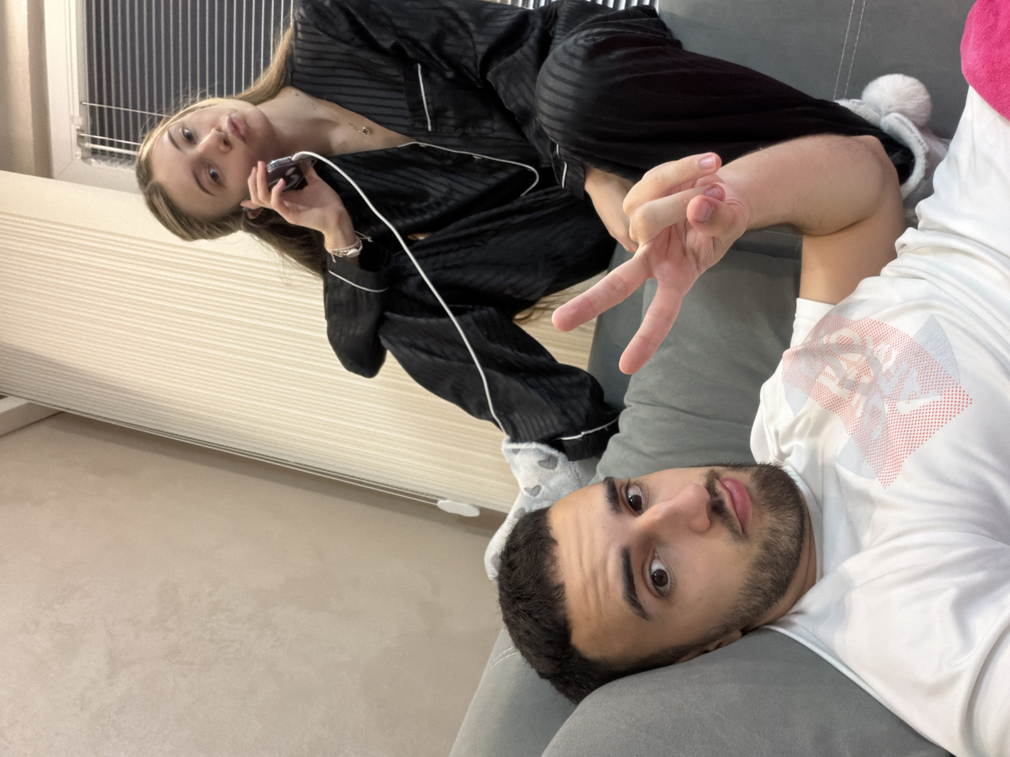 Two individuals are on a gray couch, likely indoors. A woman with long blonde hair is dressed in black striped pajamas and appears to be holding a phone to her ear. A man with short dark hair and a beard is lying on the couch, making a peace sign with his hand. He is wearing a white t-shirt. The background shows a wall and window blinds. There is no visible information to ascertain the exact location in Plovdiv, Bulgaria.