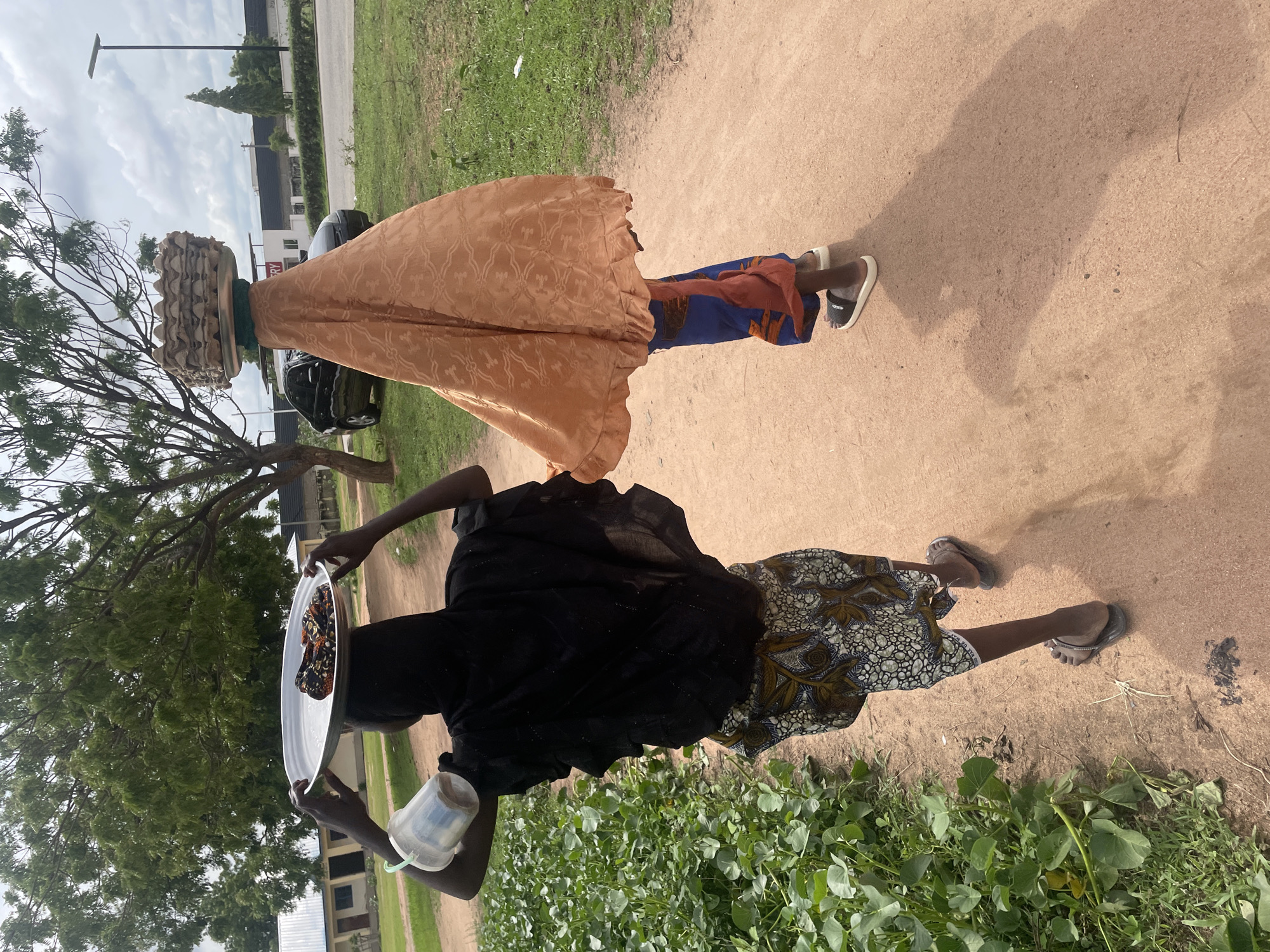Two individuals are walking on a dirt path next to lush green vegetation. The person in the foreground, facing away from the camera, is carrying a metal tray filled with food items on their head, balanced by their hands. This individual is wearing a black headscarf and a patterned skirt. A plastic cup with a green handle hangs from their left arm. The second individual, to the right and slightly ahead, is also facing away and appears to be carrying a basket of items on their head, covered by an orange cloth that drapes down to their knees. This person is wearing a blue garment and a red cloth around their waist. Both are wearing sandals. In the background, there are buildings, trees, a road with parked cars, and a street lamp under a cloudy sky. The setting appears to be an outdoor, possibly rural or semi-urban environment.