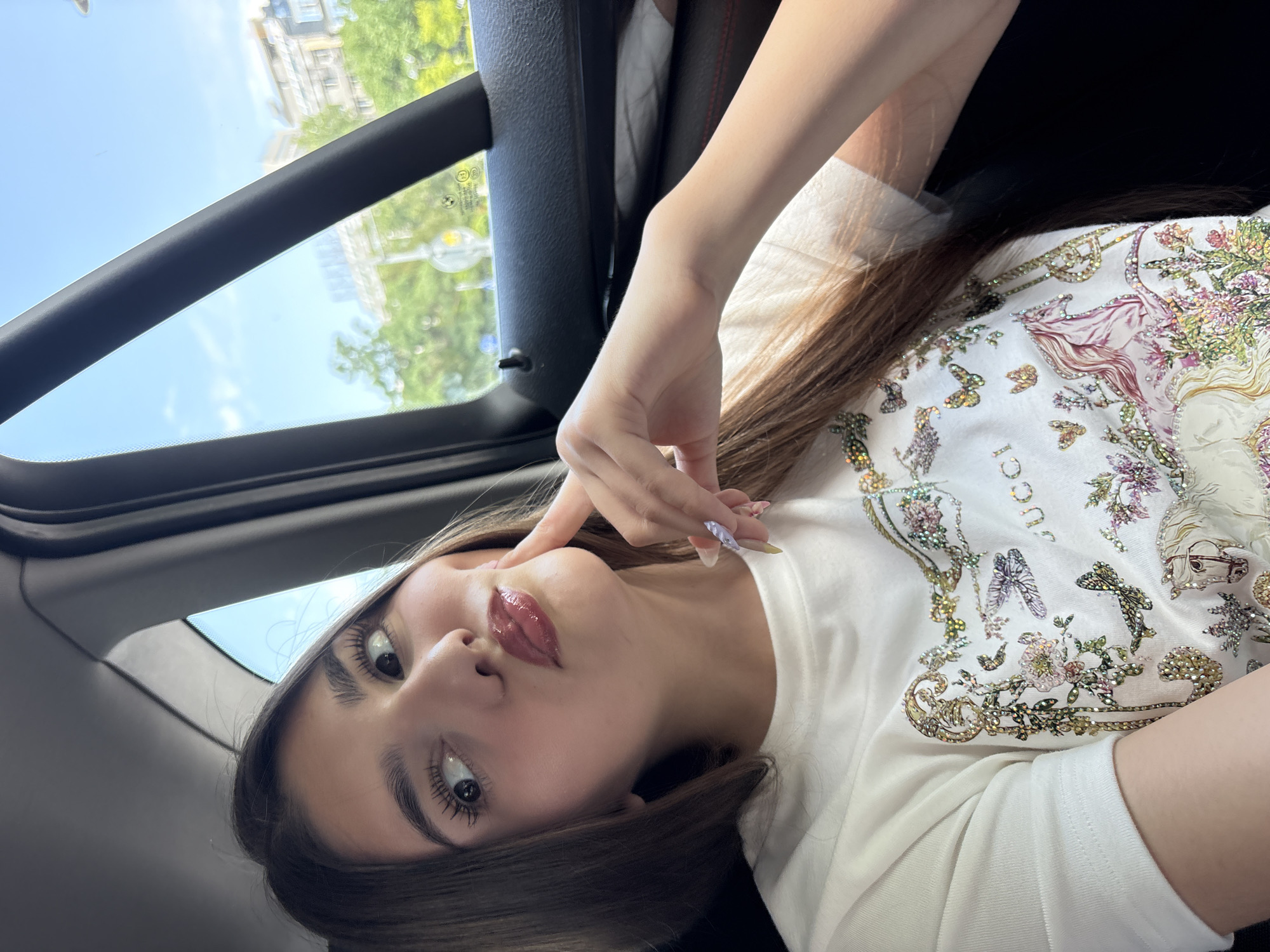 The image depicts a young adult female seated inside a vehicle, positioned to the left of the frame. She has long, straight, dark brown hair and is looking upwards and to the right with wide eyes, her lips puckered in a "duck face" expression. Her right index finger is pressed against her right cheek. She is wearing a white, short-sleeved t-shirt featuring a multi-colored, embellished design that includes the word "GUCCI," various floral patterns, butterflies, and a stylized white horse with a pink mane. Her fingernails are long, shaped, and painted with light-colored, possibly ombre, polish. The interior of the car visible includes a dark headliner, dark window frames, and a dark door panel with faint red stitching. Through the car window, a clear blue sky is visible above an urban landscape. This landscape includes green trees, several light-colored multi-story buildings, and a street scene with a pole and a circular road sign. A small BMW logo sticker is visible on the lower section of the car window. The scene is illuminated by bright daylight.