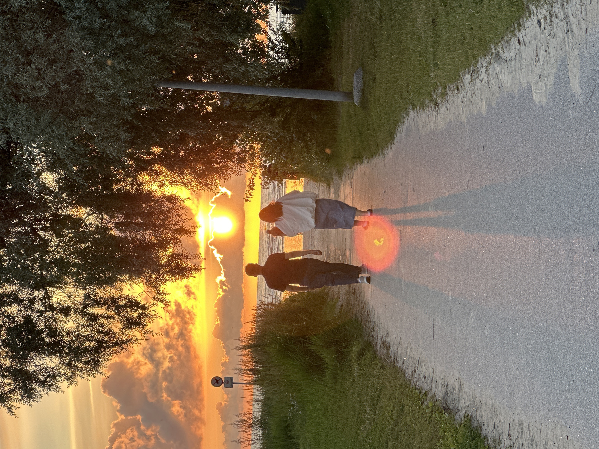 The image depicts a path at sunset in Tallinn, Estonia. A man and a woman are walking away from the camera on the paved path. The man is wearing a dark polo shirt and dark pants, while the woman is wearing a light-colored jacket over a dark skirt. They are walking towards the ocean, which is visible in the background. The sky is filled with dramatic clouds illuminated by the setting sun, creating a vibrant orange and yellow glow. Tall grass and trees line the path, and a lamppost is visible to the right of the path. The overall impression is one of a peaceful evening stroll.