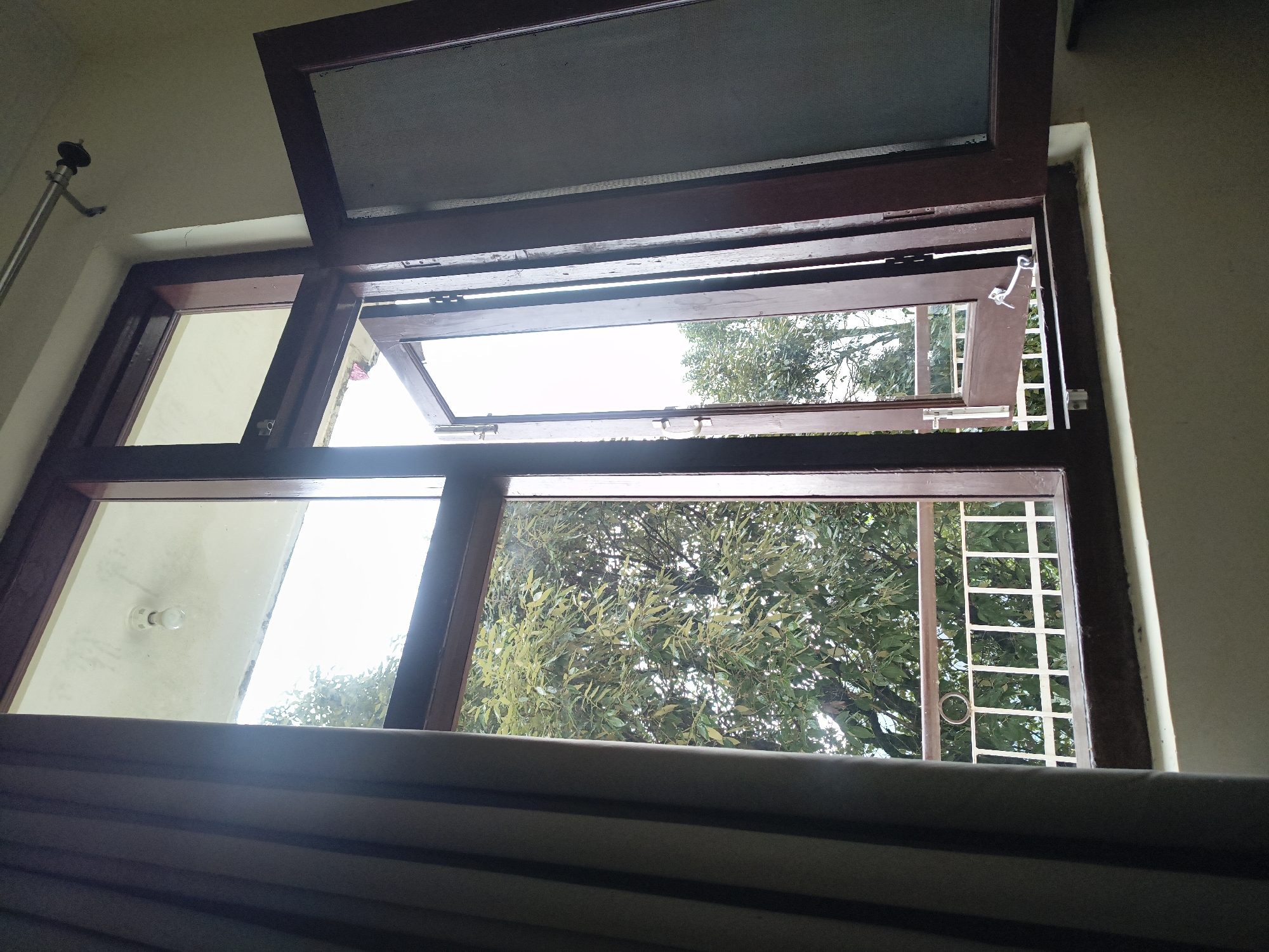 The image shows a view from inside a room looking out through a wooden-framed window. The window has multiple panes and one of the casement windows is open, revealing a balcony with a white railing and a railing post. Beyond the balcony, lush green trees with visible leaves are seen against a bright sky. A light bulb is visible on the ceiling inside the room. The scene is well-lit by natural light coming through the window. There are no people or animals in the image. The overall setting appears to be an interior space overlooking an outdoor natural environment.