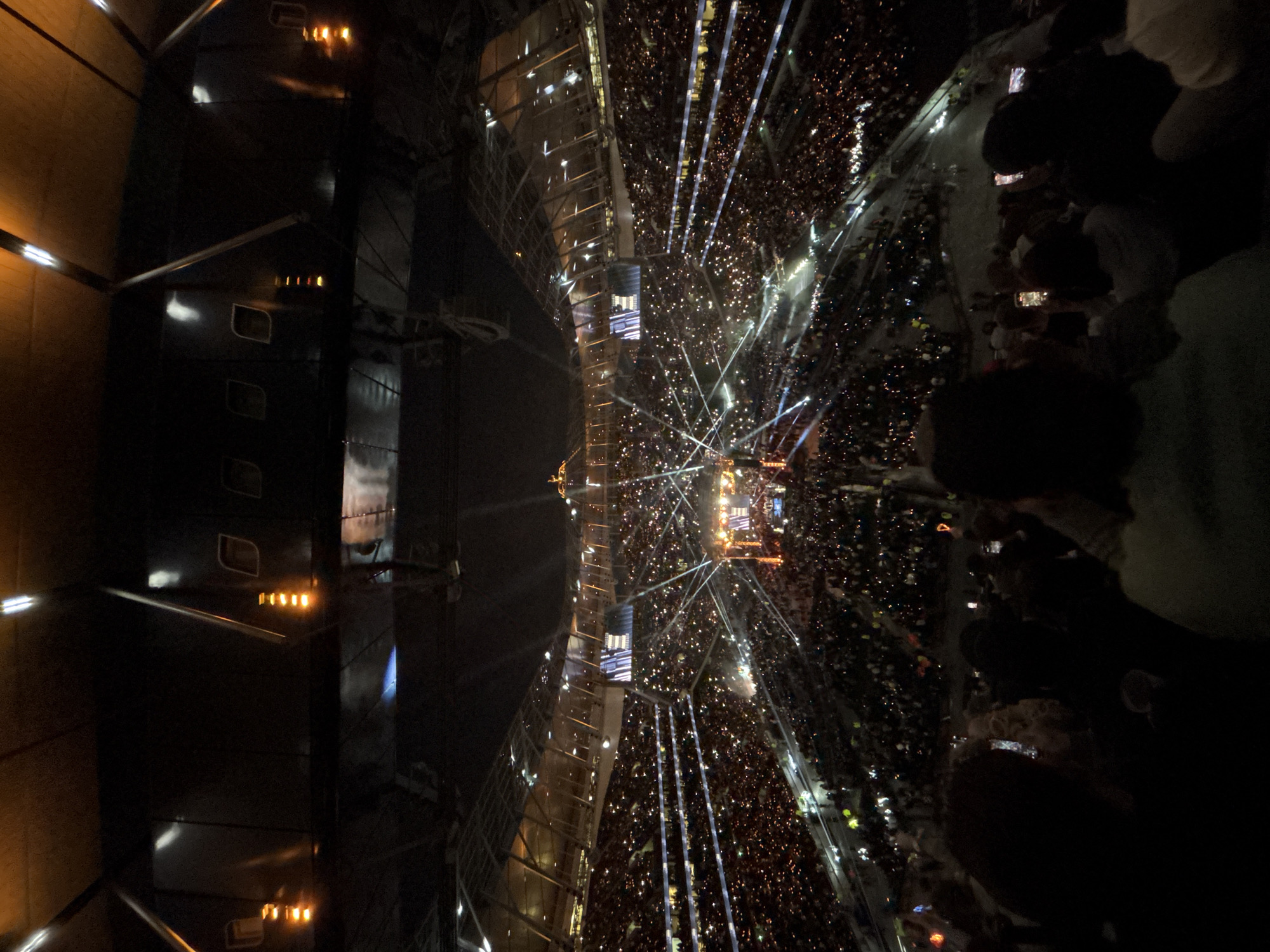 The image depicts a large stadium concert at night in London, United Kingdom. A massive crowd is gathered, illuminated by stage lights and the glow of cell phones. The stage is centrally located, with rigging and screens visible. People are standing and holding up their phones, presumably recording the event. The stadium architecture, with multiple levels of seating, is partially visible. A side structure on the left has lighted panels. People can be seen standing in the dark, likely at the perimeter of the seating.