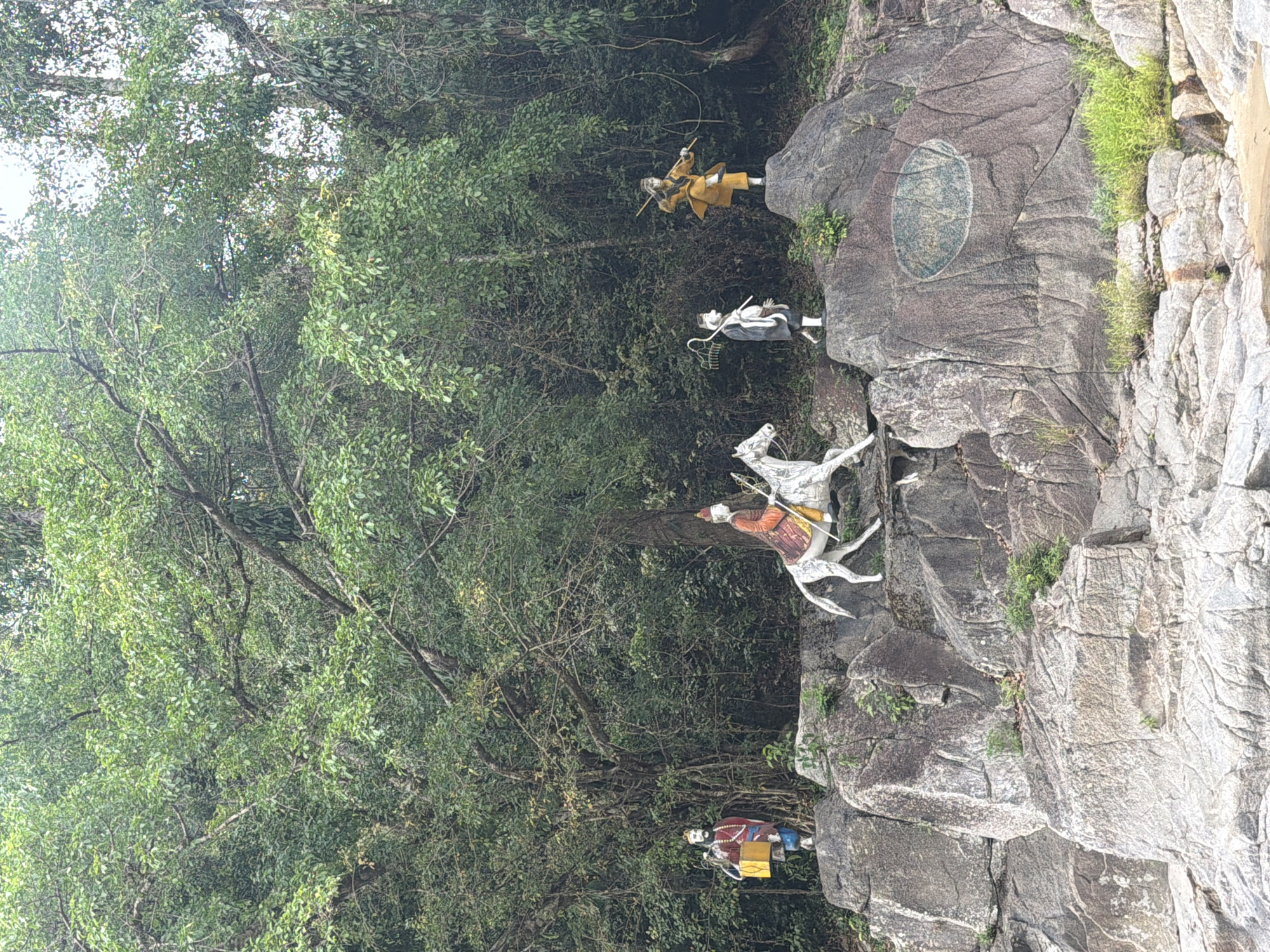 The image depicts a rocky outcrop in Khanh Vinh, Vietnam, adorned with four statues. On the far left, a statue of a man in a red and yellow tunic stands holding a yellow bucket. To his right, a statue of a person in ornate red and gold attire rides a white horse, holding reins. Next to the horse, a statue of a person in black and white robes carries a staff. The furthest statue to the right is of a person in a yellow tunic, holding two sticks. The statues are positioned against a backdrop of lush green trees and foliage. The rocky terrain is rough and uneven, with various plants growing from crevices. A large, oval-shaped carving or painting is visible on the rock face behind the statues. The foreground shows the edge of a body of water.