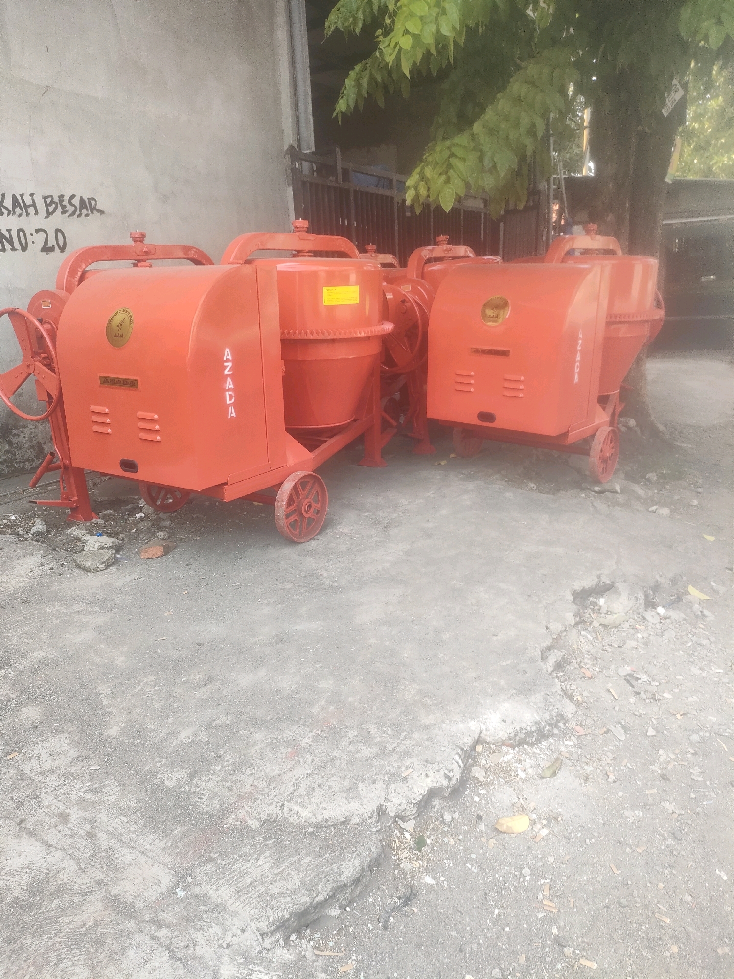 This is a medium-shot, outdoor photograph of several industrial concrete mixers, painted a bright orange. The mixers are arranged on a paved surface that appears to be a roadside or an open lot. In the background, a weathered concrete wall has graffiti on it that reads "KAH BESAR NO: 20". To the right of the mixers, a large tree with green leaves is visible. The scene suggests a commercial or industrial area in Surabaya, Indonesia, possibly a construction equipment yard or a seller's lot.