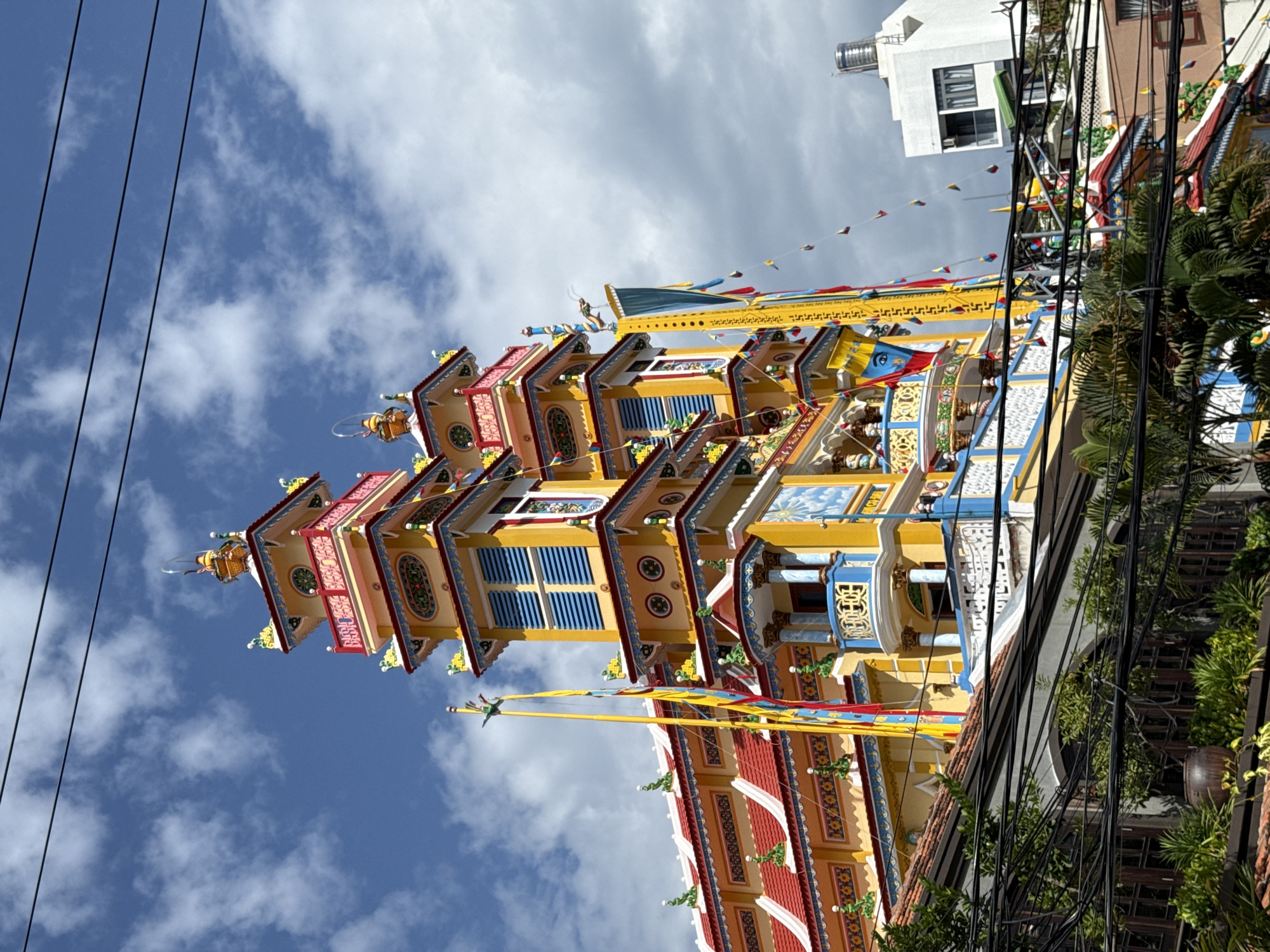 The image captures a vibrant, multi-tiered pagoda, likely a temple, with intricate architectural details. The structure is predominantly yellow and blue with red accents on the rooflines and decorative elements. Several flags and banners, in various colors including yellow, red, and blue, are strung across the building and on poles. The pagoda features multiple levels with balconies, ornate railings, and decorative motifs, including what appear to be dragon sculptures and celestial imagery. Above the pagoda, a bright blue sky with scattered white clouds is visible, along with several black electrical wires crisscrossing the frame. In the background, a modern white building with dark windows stands out. In the foreground, lush green foliage and tangled electrical wires obscure the lower portion of the scene, suggesting an urban setting. The overall impression is one of a colorful and culturally significant religious site set against a backdrop of a cloudy sky and encroaching urban elements. This pagoda is likely the Giac Lam Pagoda, a notable landmark in Nha Trang, Vietnam.