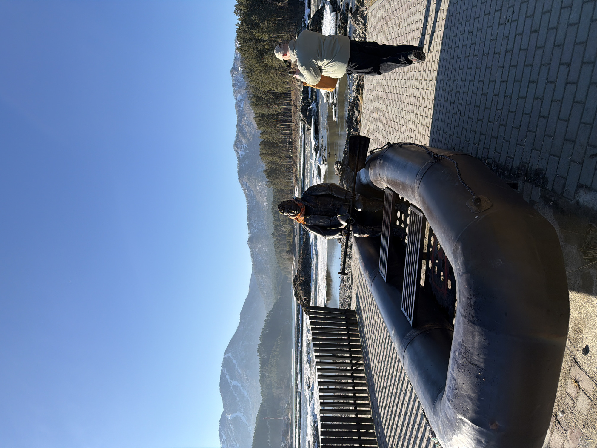 The image depicts a sunny outdoor scene in Manzherok, Russia, featuring a statue of a river raftsman on a metal contraption that resembles a raft, positioned along a brick walkway. In the background, a mountain range rises under a clear blue sky. A body of water, possibly a river, is visible with ice floes. A person wearing a light-colored jacket and dark pants is standing on the walkway, appearing to take a photograph. The scene is captured in a landscape orientation.