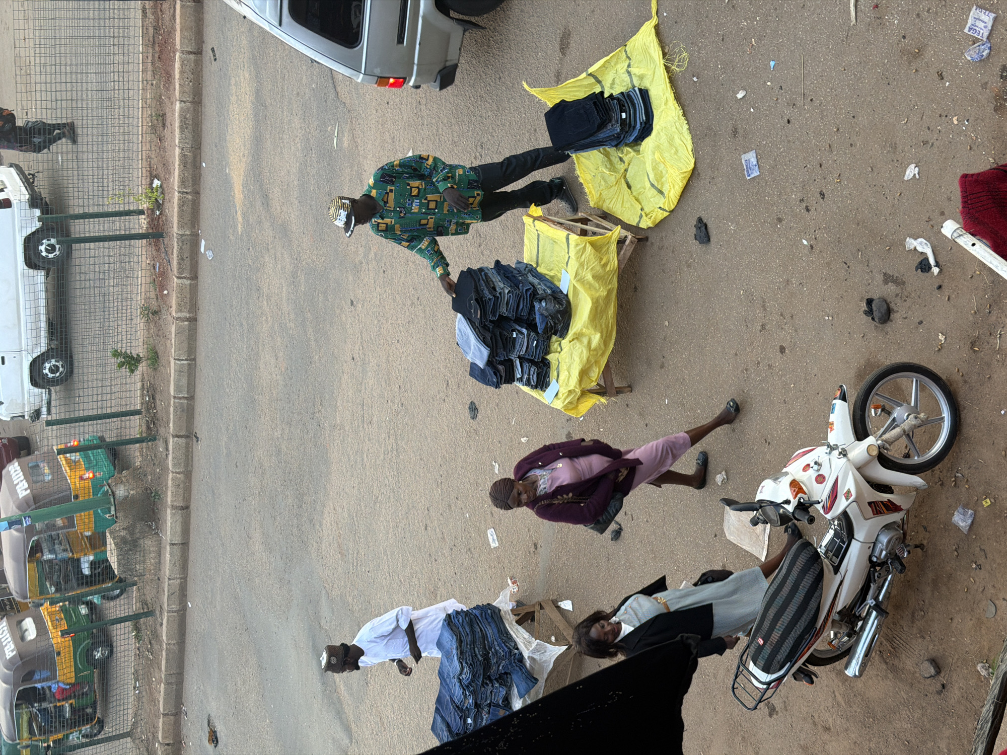 A medium shot captures a bustling street scene in Bukuru, Nigeria, focusing on vendors and shoppers. Two men and two women are engaged in commerce. One man, wearing a white tunic and cap, stands near a stack of folded blue jeans. Another man, dressed in a colorful shirt and cap, attends to a display of folded jeans on a yellow tarp. A woman in a pink dress and purple jacket browses the merchandise, while a second woman in a black blazer and light blue dress walks past. A white motorcycle with a black and striped seat is parked in the foreground. In the background, a silver van is visible, and a green and yellow tricycle, commonly known as an "Okada" or "Keke Napep," is parked. The ground is paved with asphalt and scattered with debris.