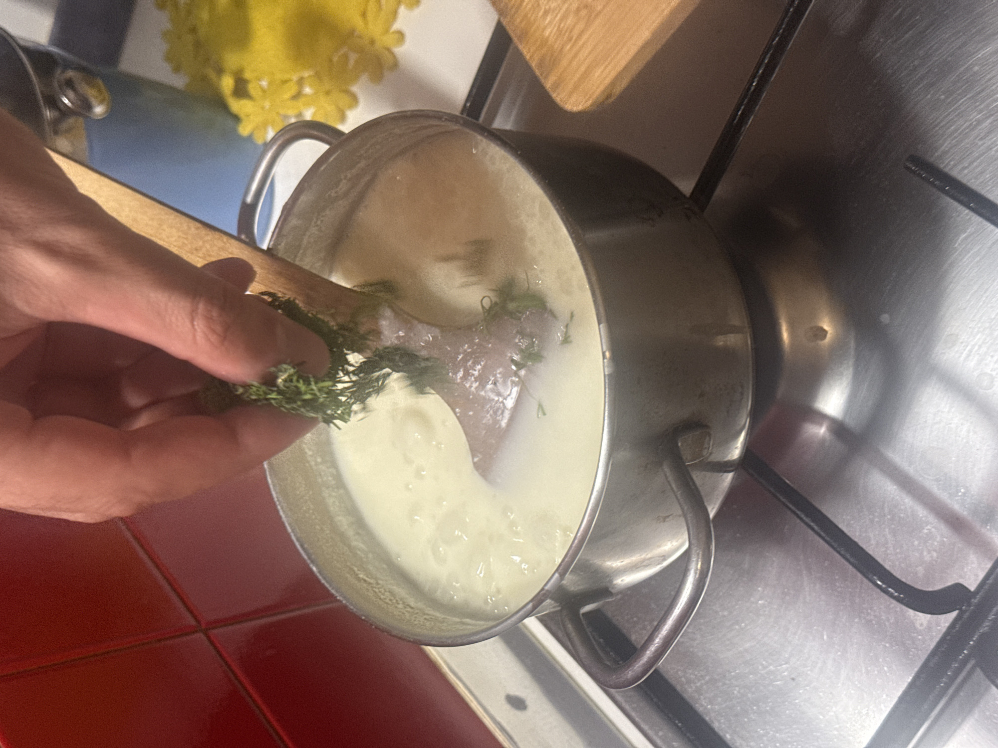 A person's hand is adding herbs to a saucepan on a stovetop. The saucepan contains a milky-looking liquid. The background shows red tiles and a stove grate. The location cannot be determined from the image.