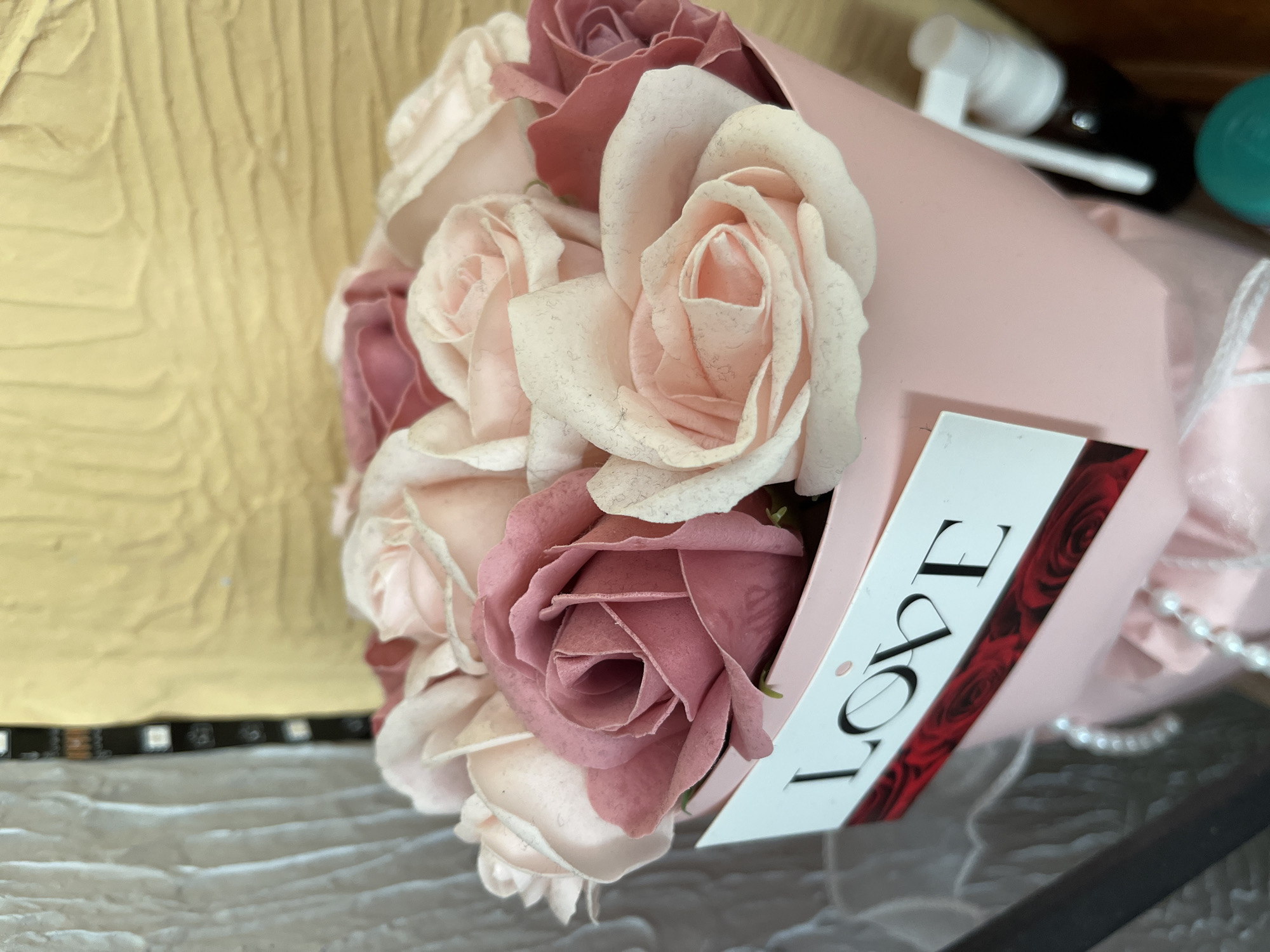 A close-up, eye-level shot shows a bouquet of artificial roses in shades of pink and dusty rose, arranged in a pale pink cylindrical holder. A white card with the word "LOVE" printed in black, bold sans-serif font is attached to the front of the holder, with a border of red roses underneath. The bouquet is positioned in the foreground, with a textured yellow wall and a vertical black and white striped object in the background. The scene suggests an indoor setting, possibly a gift shop or a home, in Kalynivka, Ukraine. There are no people visible in the image.