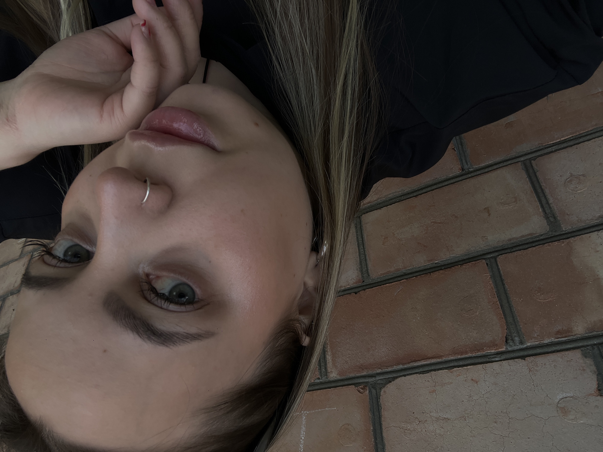 This media file is a close-up, angled portrait of a young person, likely female, with fair skin, light green or blue eyes, and long, straight hair with blonde highlights. The individual is wearing a dark, possibly black, top or jacket. Notable accessories include a silver hoop nose ring on their right nostril and a small silver earring on their left ear. One hand, with visible red nail polish on at least one finger, is partially resting near their chin and mouth. The person's lips are slightly parted, and they are looking directly at the camera with a neutral expression. The background consists of a textured brick wall with visible mortar lines, suggesting an indoor or outdoor setting against a building. The image is oriented with the subject's head tilted significantly to the left. No text is visible in the image. The location context provided is Novopokrovskaya, Russia.
