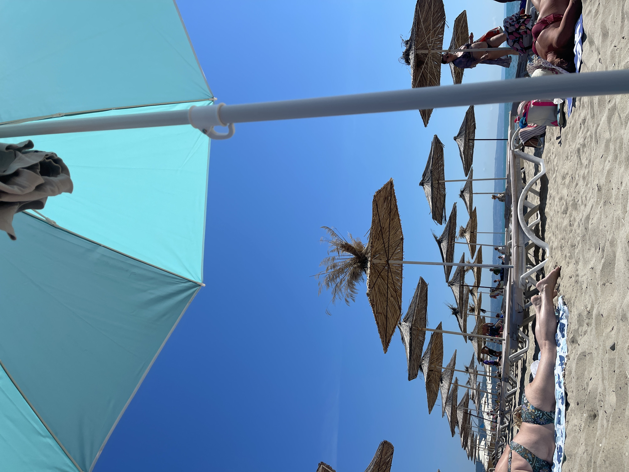 This image captures a sunny day on a sandy beach in Kranevo, Bulgaria. In the foreground, a large, turquoise umbrella dominates the frame, casting a shadow over the beach. To the left, a person in a patterned bikini lies on a towel. In the middle ground, rows of straw umbrellas and white lounge chairs stretch towards the sea, providing shade for beachgoers. Several people are visible lounging on the chairs or walking along the shore. The clear blue sky and calm sea suggest a typical summer day at the resort.