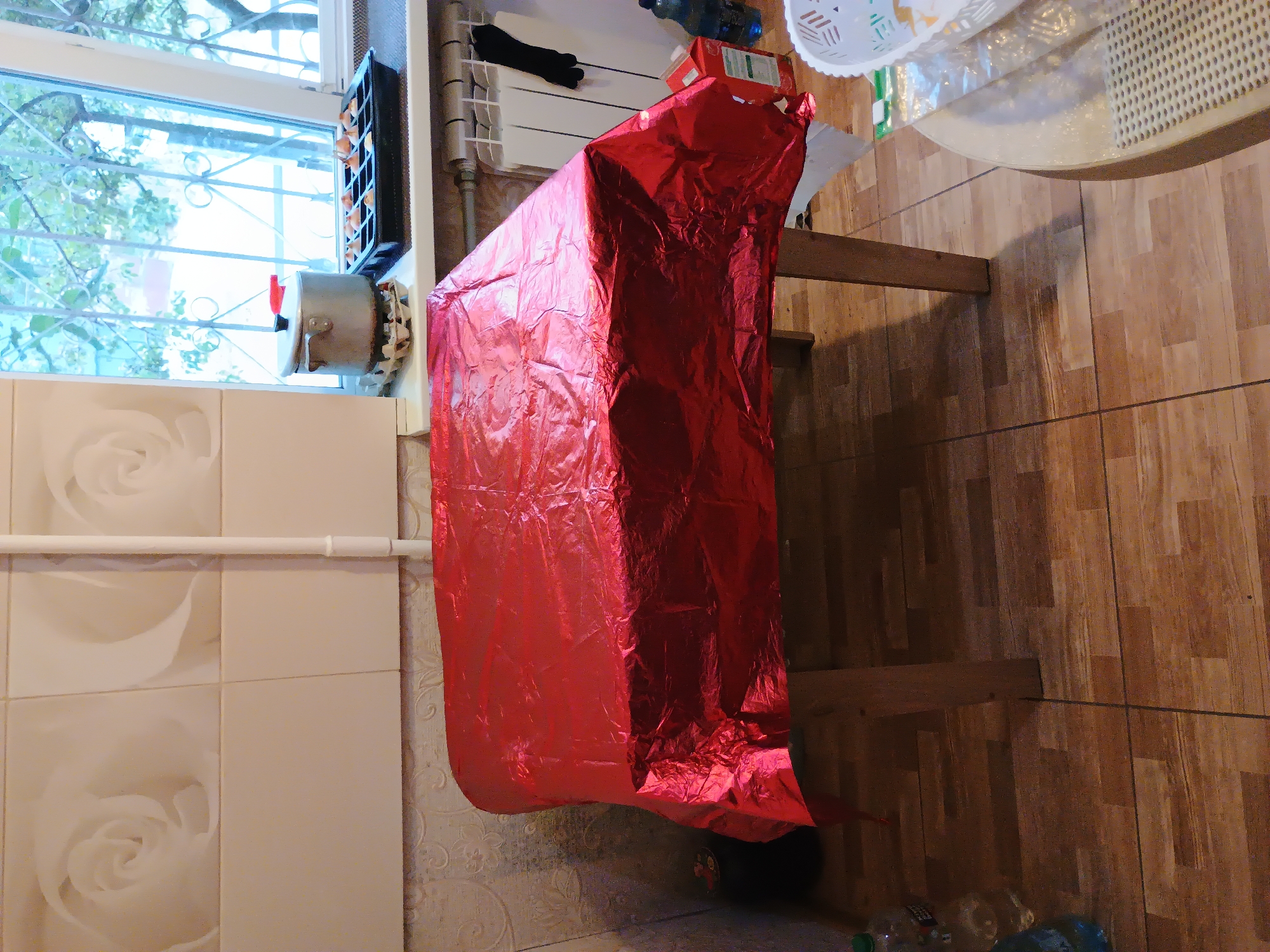 The image depicts a close-up interior view of a room, likely a kitchen, in Zaporizhzhia, Ukraine. The foreground is dominated by a table covered with a crinkled red metallic tablecloth. The table legs are visible, made of wood. To the right, a round table with a textured surface is partially visible, with a white basket on top. In the background, a window with a decorative metal grille shows trees and foliage outside, suggesting it's daytime. On the windowsill, a pot with a lid and a tray containing small seedlings or bulbs are present. The wall behind the table is tiled with a floral pattern. A white vertical pipe runs along the wall. Further to the right, a white radiator is visible, with a black glove hanging from it. Various bottles and a red carton are on the floor near the radiator. The floor is tiled with a wood-like pattern.
