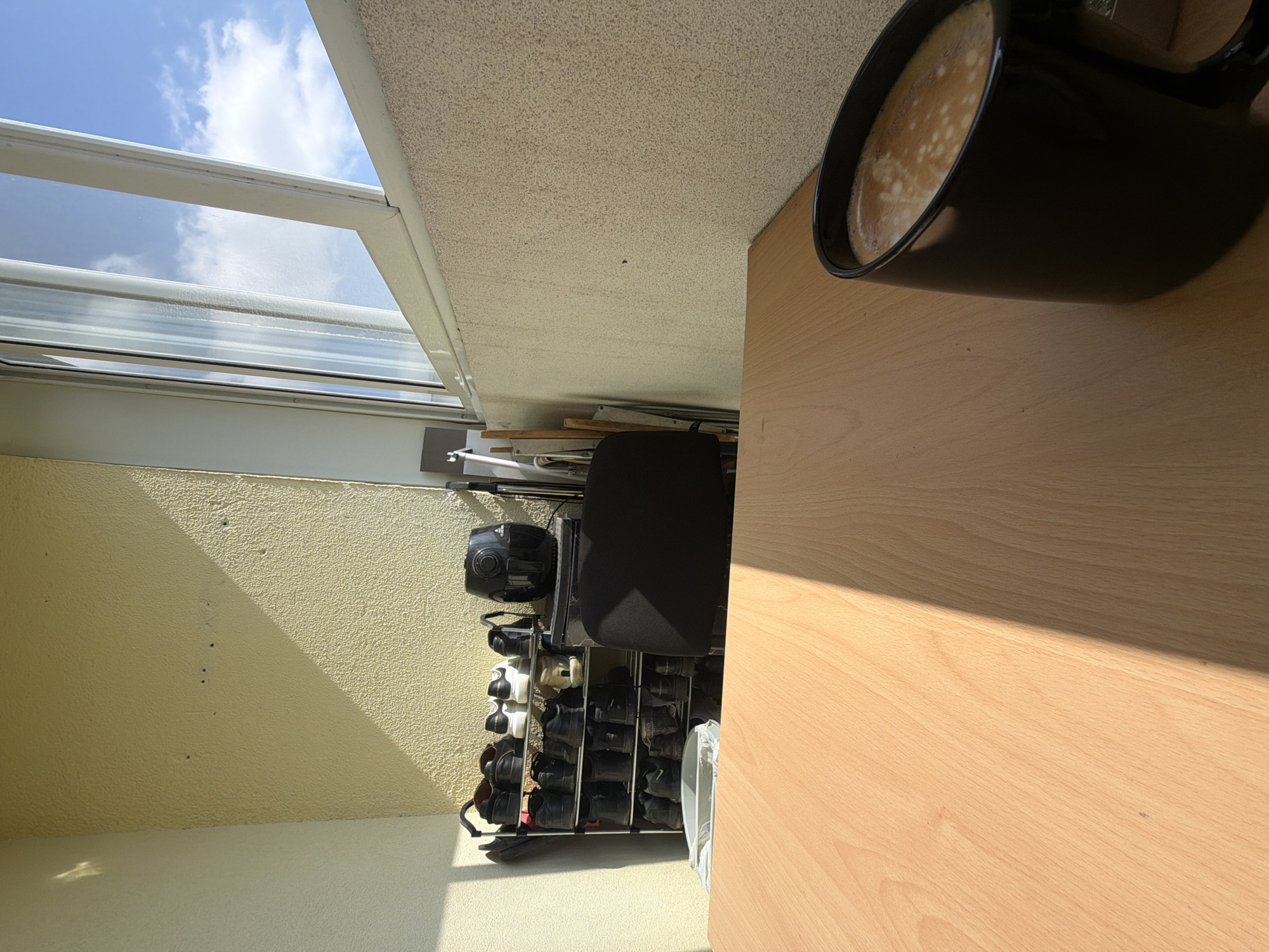 This media file presents a bright, sunlit domestic scene, likely a balcony or terrace view, captured from a tilted perspective. The upper section of the image reveals a clear blue sky adorned with fluffy white clouds, framed by a window or a transparent roof structure, suggesting an enclosed or semi-enclosed outdoor space.
On the left, a textured, light yellow or cream-colored wall is illuminated by direct sunlight, casting a distinct diagonal shadow across its surface. In the foreground on the right, a light-toned wooden table or desk is visible, on which a black mug containing a frothy, light brown beverage, possibly coffee, is placed. The mug and an unseen object cast sharp, elongated shadows across the wooden surface, indicating bright sunshine.
Below the yellow wall and to the left of the table, a metal shoe rack neatly stores numerous pairs of shoes, predominantly dark in color, with a few lighter ones. A black office chair is positioned next to the shoe rack, and a black appliance, possibly an air purifier or a speaker, rests above the shoes. Miscellaneous items, including stacked wooden planks and metal rods, are visible behind the furniture.
The scene captures a peaceful daytime moment, likely morning or afternoon, given the intensity and angle of the shadows. The setting appears to be a residential area in Sofia, Bulgaria. No people or visible text are present in the image.