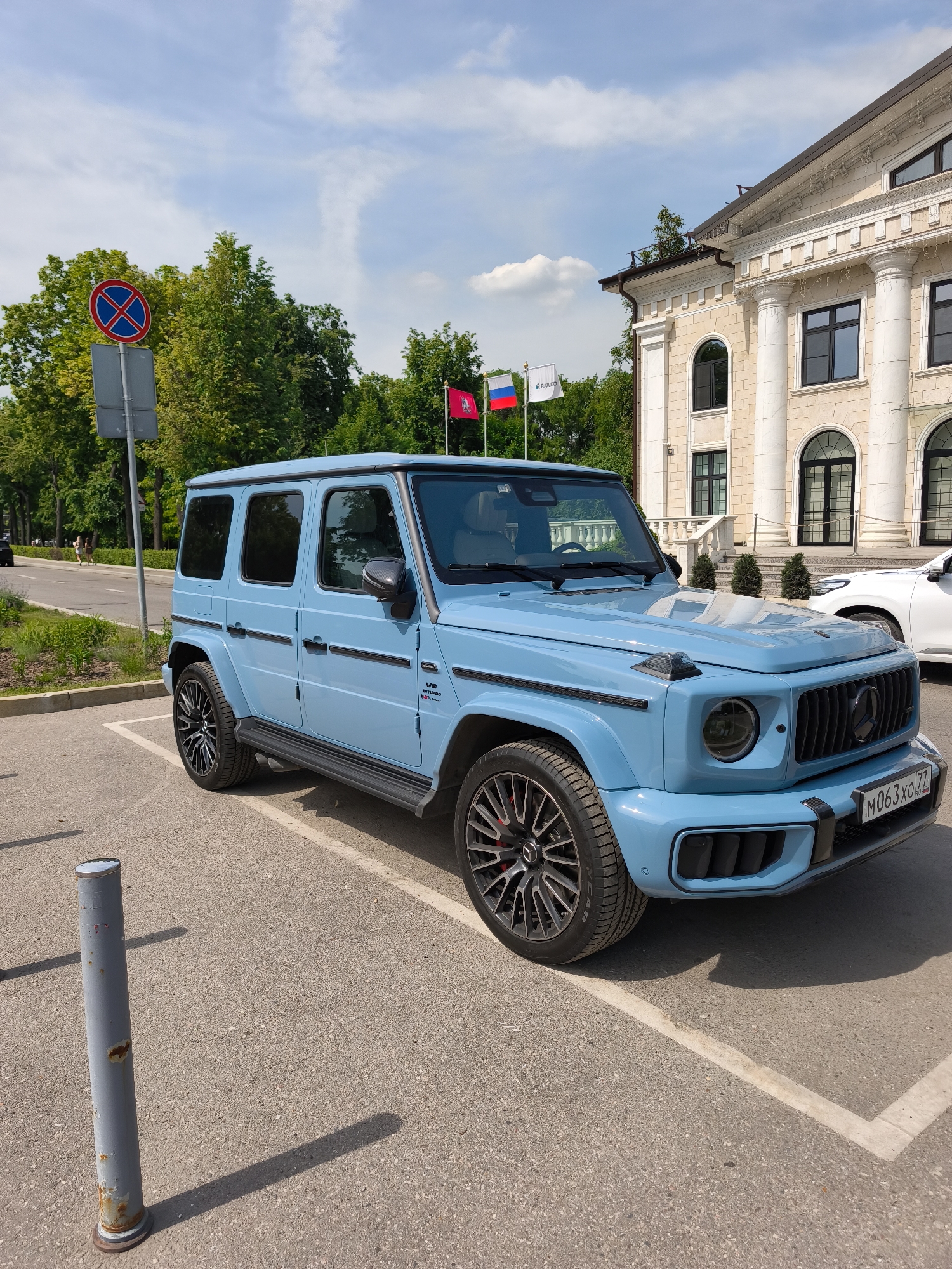 The image captures a light blue Mercedes-Benz G63 AMG, identified by its grille, wheels, and "V8 BITURBO" and "G Manufaktur" badging on the side. The car is parked on a clear asphalt surface with white parking lines, positioned with its front facing right and slightly angled towards the camera. The license plate "M063XO 77 RUS" is visible on the front bumper. The vehicle features dark multi-spoke wheels with red brake calipers, carbon fiber-look side mirrors, and black running boards.
In the background, a grand, multi-story building with neoclassical architecture stands prominently. It is constructed from light-colored stone or stucco, featuring large classical columns, ornate cornices, and balustrades. The building has a mix of arched windows on its lower level and rectangular windows on the upper floors, all with dark frames. This structure is identified as the Moscow Railway Management building at Komsomolskaya Square, 3/3, Moscow, Russia.
Three flags are visible flying from poles in front of the building. From left to right, these are the national flag of the Russian Federation (white, blue, and red horizontal stripes), a white flag bearing the black "RAILCO" logo, and a red flag displaying the golden emblem of Saint George slaying the dragon, which is the Coat of Arms of Moscow.
To the left of the car, a red circular "No Stopping" or "No Parking" road sign (a blue circle with a red border and a diagonal red cross) is visible on a grey pole. Green deciduous trees line a paved road in the mid-ground, beyond which two indistinct figures appear to be walking on a sidewalk. A white vehicle is partially visible parked behind the blue G-Wagen, closer to the building's entrance. In the immediate foreground, a grey, slightly corroded metal bollard casts a long shadow to its right. The scene is brightly lit by sunlight under a clear blue sky with sparse white clouds.