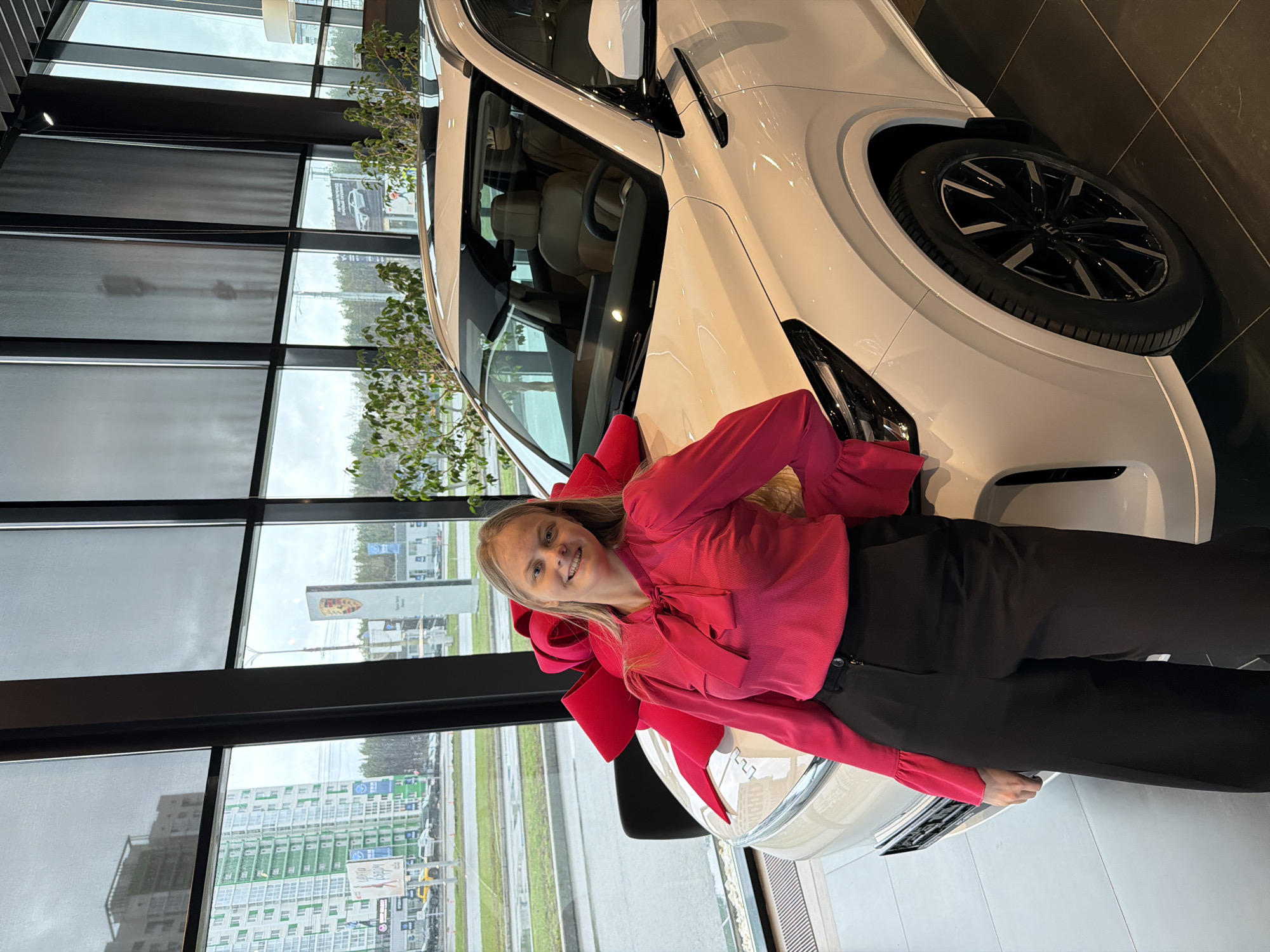 A medium shot depicts a woman leaning against the hood of a white car adorned with a large red bow, likely a gift. The woman has fair skin and light brown hair, wearing a bright pink long-sleeved shirt and black pants. She is smiling towards the camera. The car is parked inside what appears to be a car dealership with a large window overlooking a city landscape. Through the window, buildings and billboards are visible; one billboard seems to show a Porsche logo, suggesting the location is a dealership for that brand. Based on the available signage, the location is potentially identifiable as Izhevsk, Russia. The interior of the dealership features dark window frames and a tiled floor, creating a modern and polished environment.