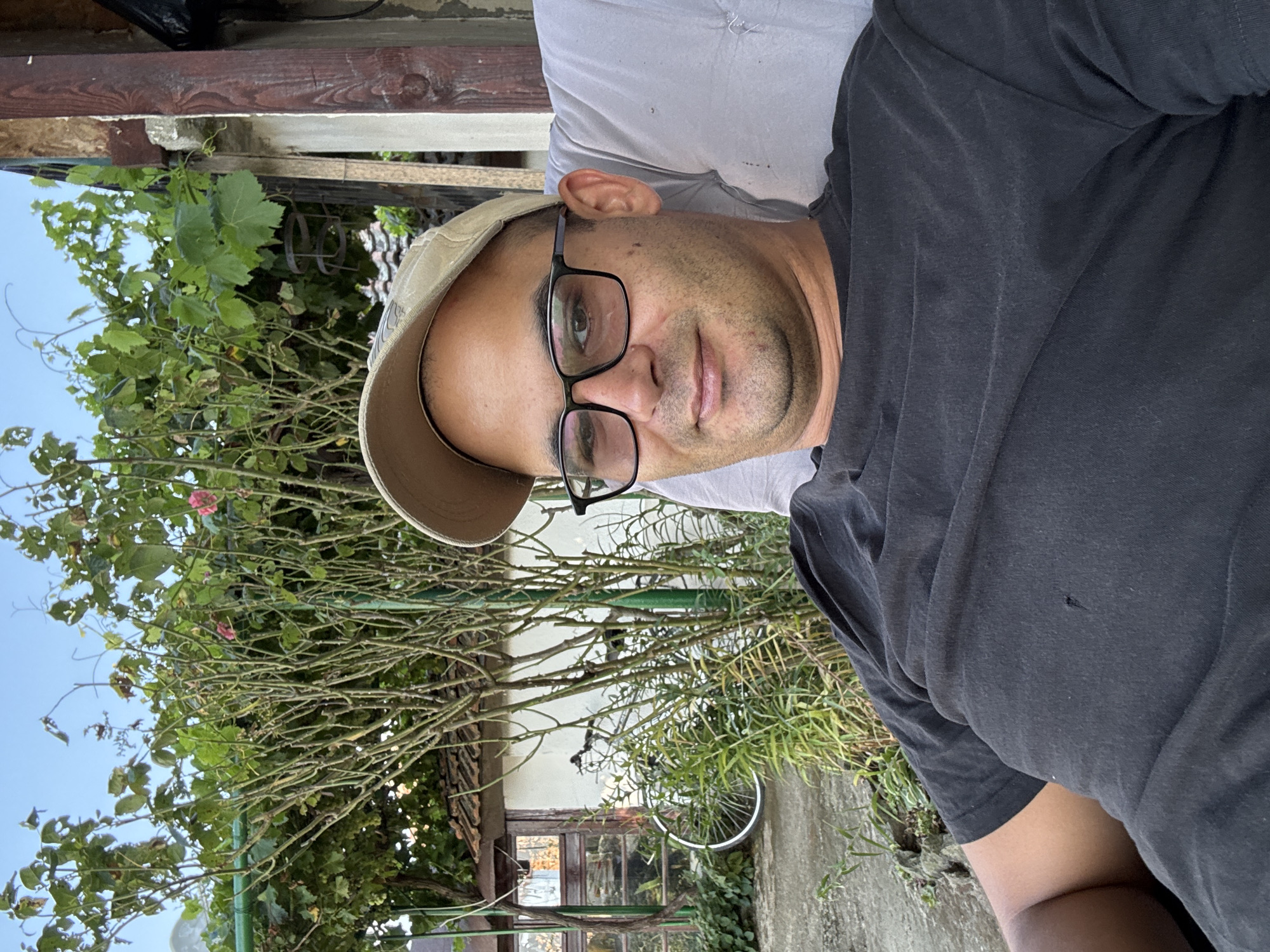 A man with glasses and a baseball cap is seated outdoors, looking at the camera. He is wearing a black t-shirt. Behind him, there is lush greenery, including a vine with leaves and some pink flowers. A portion of a building with a tiled roof and wooden beams is visible. A bicycle wheel is also present in the background. The sky is clear and blue. The setting appears to be a residential yard, possibly in Generalovo, Bulgaria, given the architectural style of the building and the vegetation.