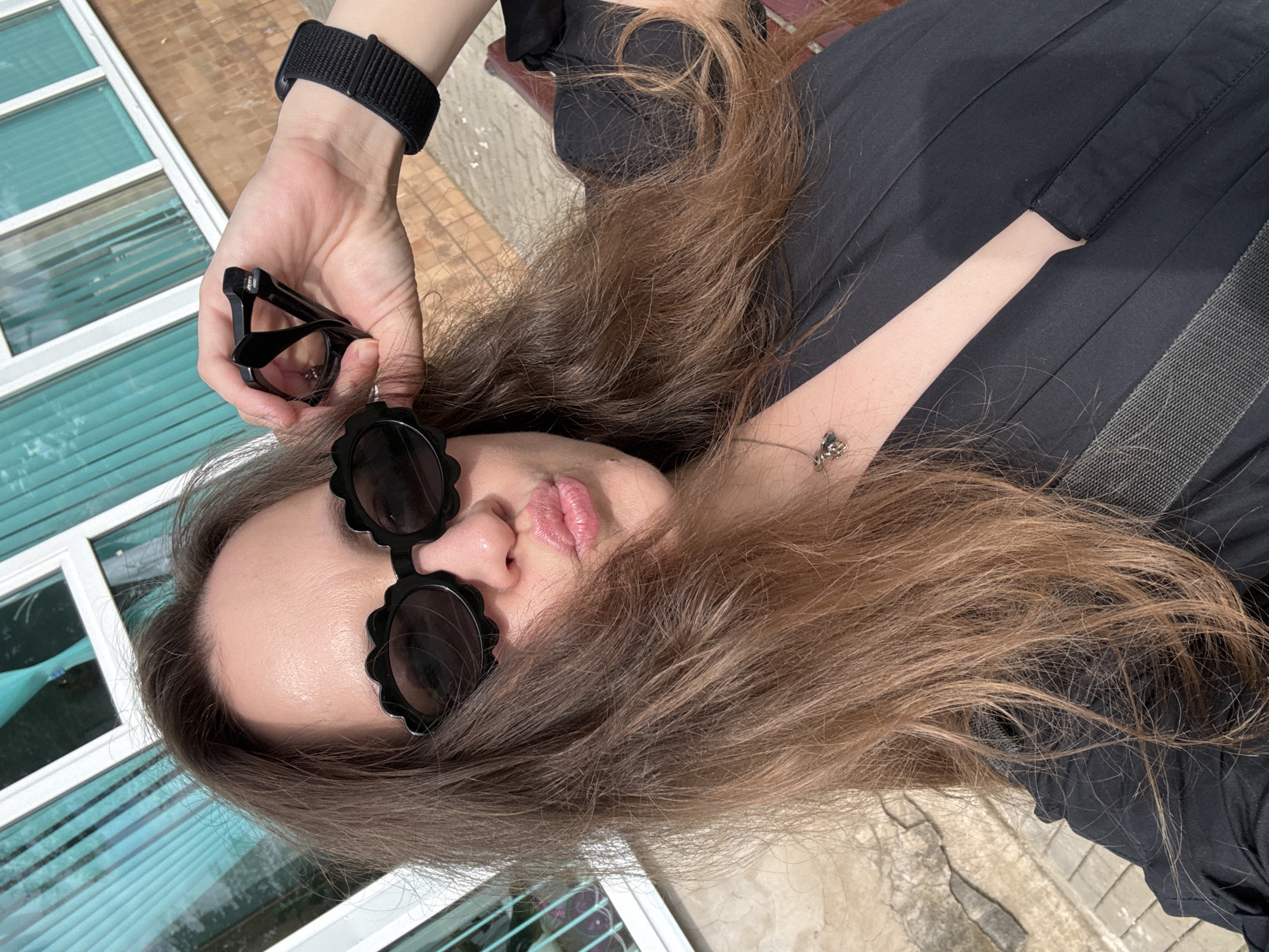 The media file shows a close-up, overhead shot of a person lying down outdoors. The person has long, light brown hair that is spread out. They are wearing a black top. In their right hand, which is held up, they are holding a pair of black glasses. They are also wearing black, flower-shaped sunglasses. The person's face is partially visible, with their lips slightly parted. They are wearing a black watch on their left wrist. The background shows a light-colored building with windows, and some cracked pavement. The lighting suggests it is daytime, and the weather appears to be clear. The overall mood is casual and relaxed.