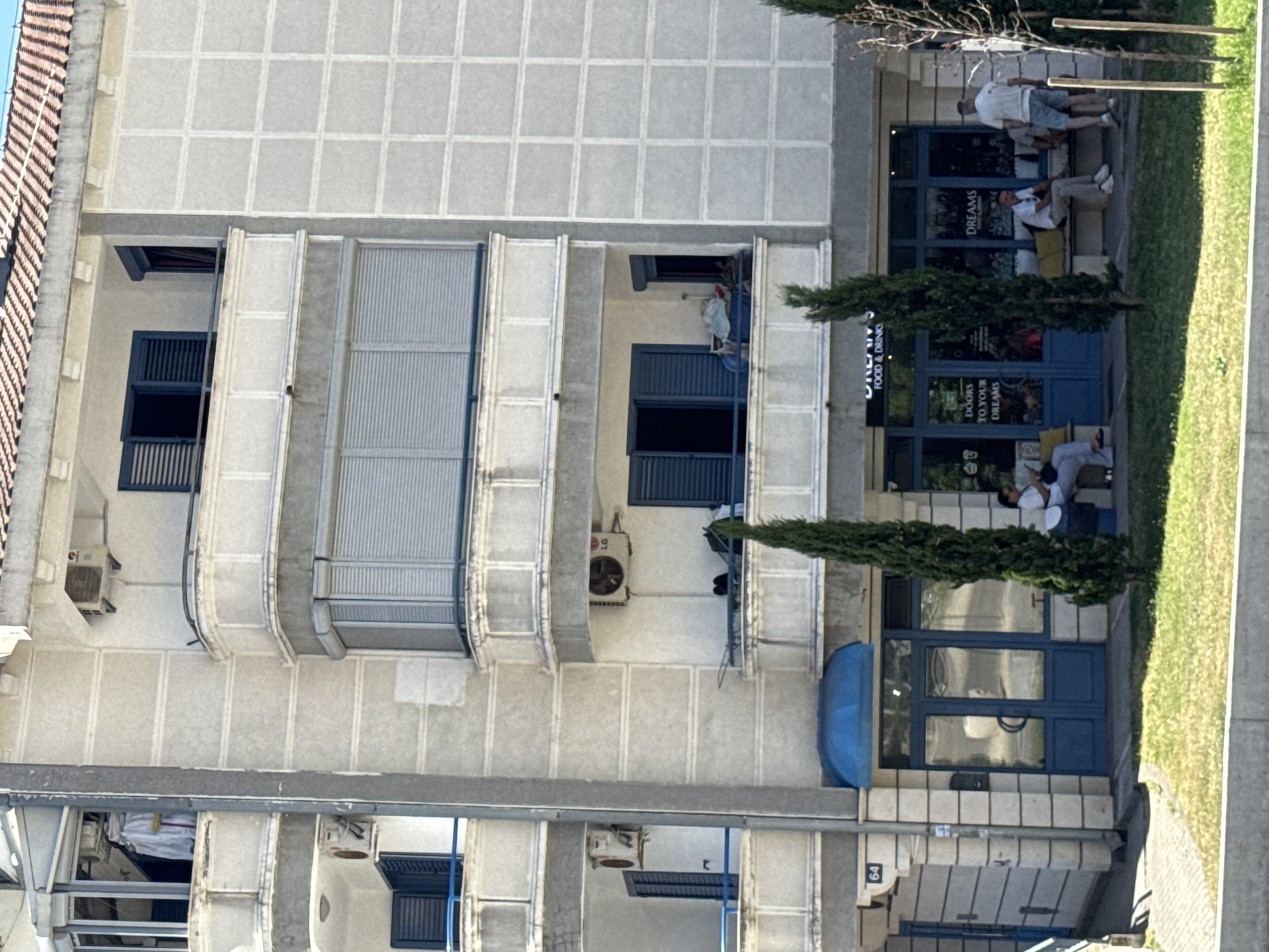 This is a vertical, eye-level shot of the exterior of a building with a restaurant or cafe. The building's facade is made of light-colored, textured panels arranged in a grid pattern. Several balconies with metal railings are visible, some with closed shutters.
In the foreground, a blue, canoe-like object is leaning against the building. To the right, a paved pathway runs alongside a grassy area.
At street level, there are blue-framed glass doors and windows, suggesting a commercial establishment. Text on the doors reads "DREAMS FOOD & DRINK" and "DOORS TO YOUR DREAMS".
Several people are seated outside the establishment on a low wall or bench, some interacting with their phones. Two individuals are standing near the entrance. The lighting suggests it is daytime, likely late afternoon due to the warm sunlight casting shadows. The weather appears to be fair. The presence of the word "Budva" in the location context suggests this scene is in Budva, Montenegro.