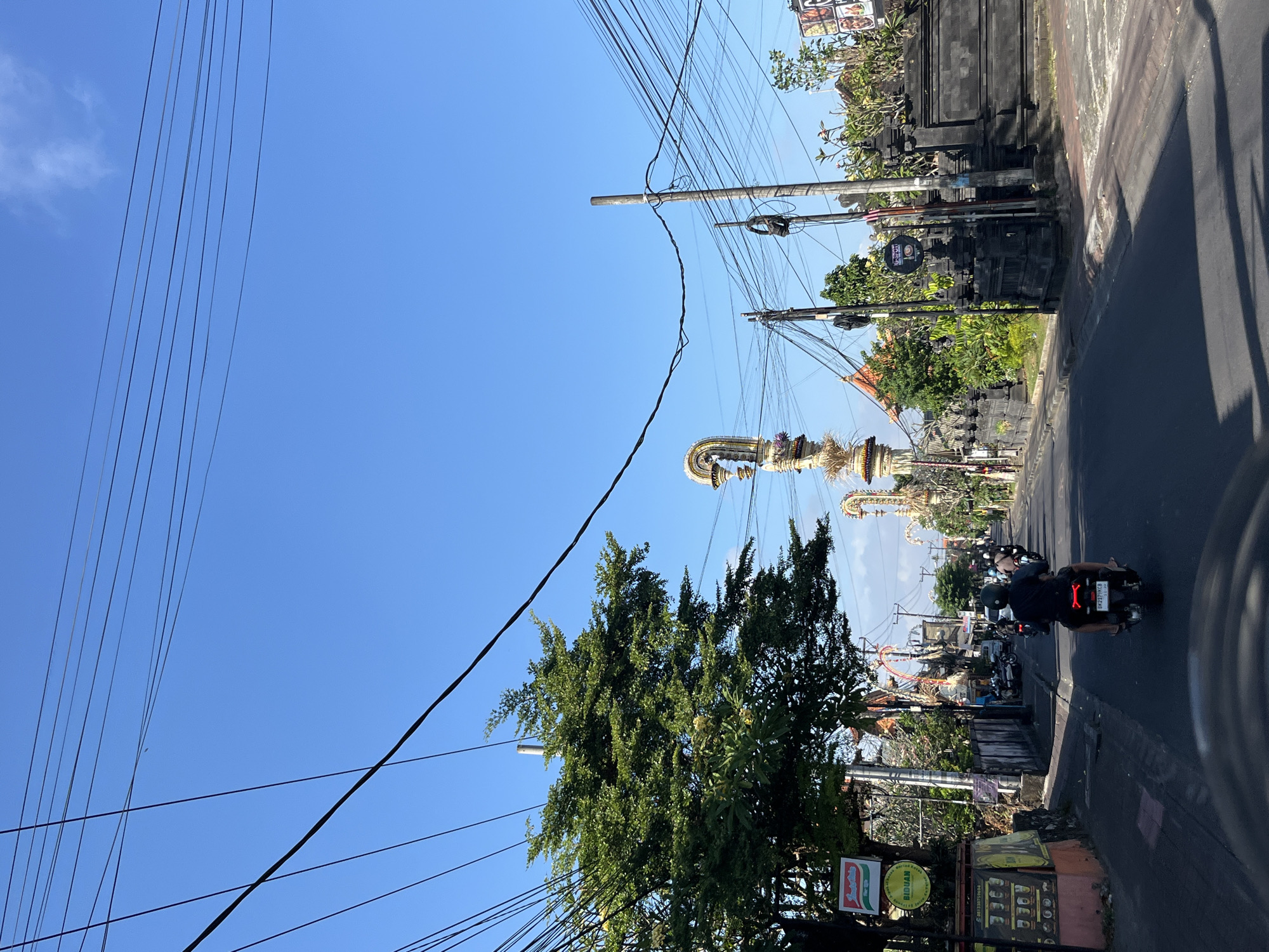 A street scene in North Kuta, Indonesia, shows a road with several motorcycles driving on it under a clear blue sky. The road is flanked by buildings, some with decorative stone walls and traditional Balinese architecture. Elaborate golden structures, possibly religious or ceremonial, are visible in the distance. A large tree with green foliage is on the left side of the road, and a dense network of overhead electrical wires crisscrosses the sky. The scene appears to be a typical daytime street view in a populated area.